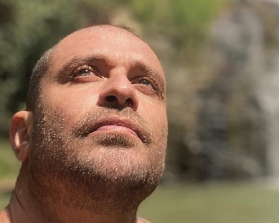 Homem de barba rala, cabeça raspada e pele molhada, olhando para cima com expressão serena, em ambiente natural com vegetação e água turva ao fundo