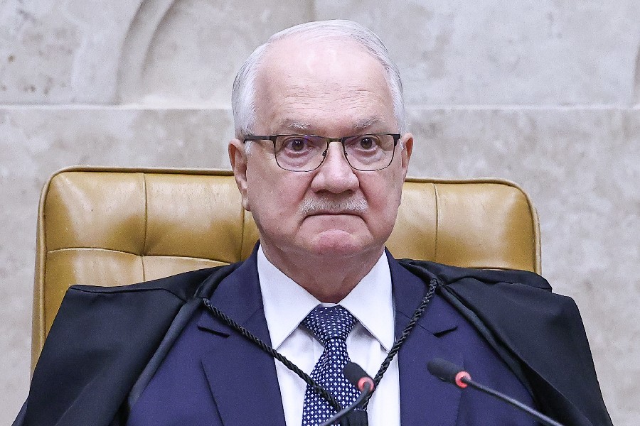 Homem idoso de cabelos brancos, óculos e bigode, veste terno azul, camisa branca, gravata azul de bolinhas brancas e toga preta, sentado em cadeira de couro marrom, com microfones à frente