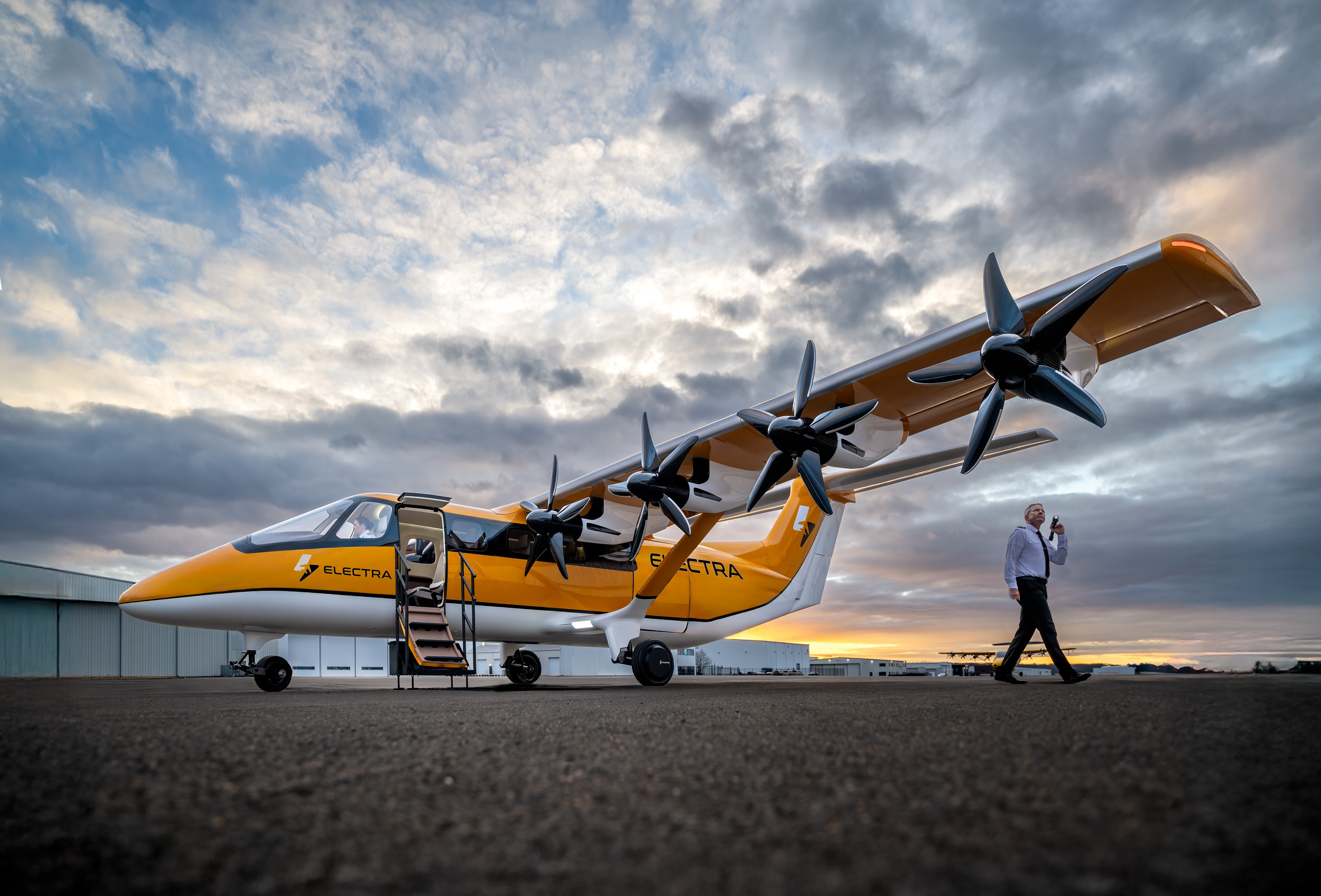 Aeronave Electra amarela e branca com oito hélices pretas na asa, porta aberta com escada, estacionada em pista de asfalto. Um homem de camisa social e calça escura caminha à direita, segurando um celular. Céu nublado com tons alaranjados ao fundo