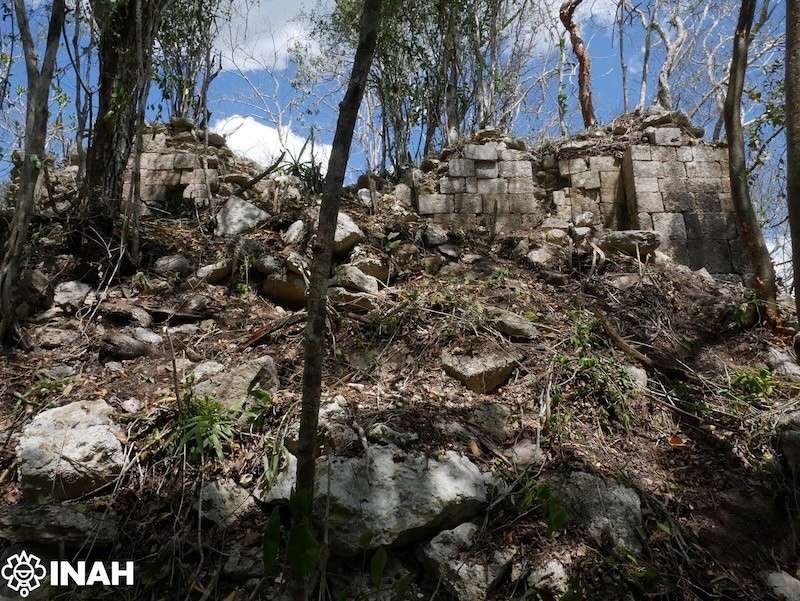 Ruínas de uma estrutura antiga de pedra, parcialmente coberta por vegetação densa e árvores, sob um céu azul com nuvens brancas