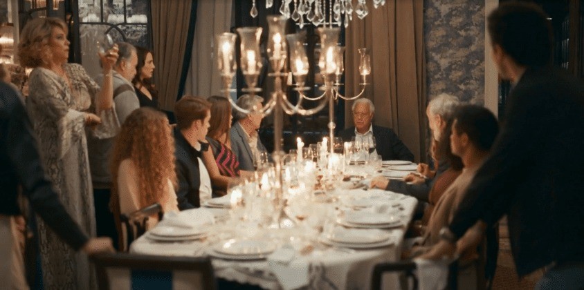 Pessoas em um jantar formal, com uma mulher loira de vestido claro em pé, erguendo uma taça. Outros convidados sentados à mesa decorada com um candelabro dourado e velas acesas, pratos brancos e taças de cristal. Um homem de terno escuro está de costas no canto direito, e um homem mais velho de terno escuro está sentado à direita da mesa. O ambiente é elegante, com cortinas e paredes escuras