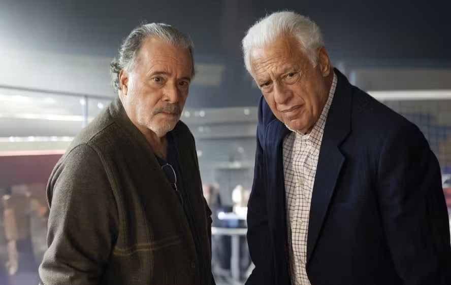 Dois homens idosos, um com cabelo grisalho e casaco marrom, outro com cabelo branco e terno azul, olham para a câmera com expressões sérias