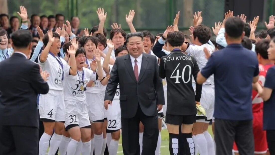 Kim Jong-un sorridente, de terno escuro e gravata vermelha, caminha entre jogadoras de futebol uniformizadas de branco, que o aplaudem com as mãos levantadas e expressões de euforia