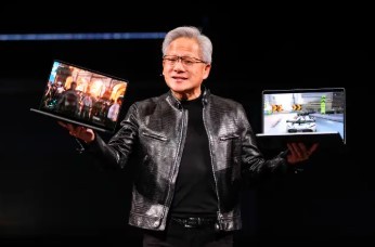 Jensen Huang, CEO da Nvidia, sorri enquanto segura um notebook aberto em cada mão, exibindo imagens diferentes nas telas. Ele veste uma jaqueta de couro preta sobre uma camiseta escura, em um palco com fundo preto.