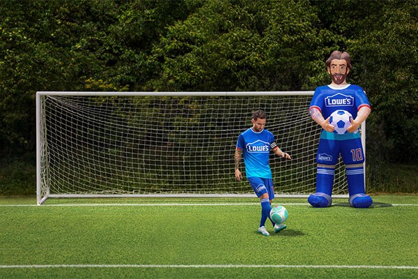 Um jogador de futebol em uniforme azul e degradê, com o logo Lowes, dribla uma bola verde e branca em um campo gramado. Ao lado, um boneco inflável gigante, também com o logo Lowes e o número 10, segura uma bola. Ao fundo, um gol e árvores.