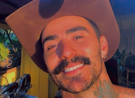 Homem sorridente com chapéu marrom, barba e bigode escuros, brincos de argola e tatuagens no pescoço, em ambiente externo com luz solar
