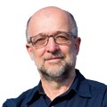 Homem branco de meia-idade, careca e com barba grisalha, usando óculos de armação escura e camisa azul-marinho, olhando para frente com expressão séria