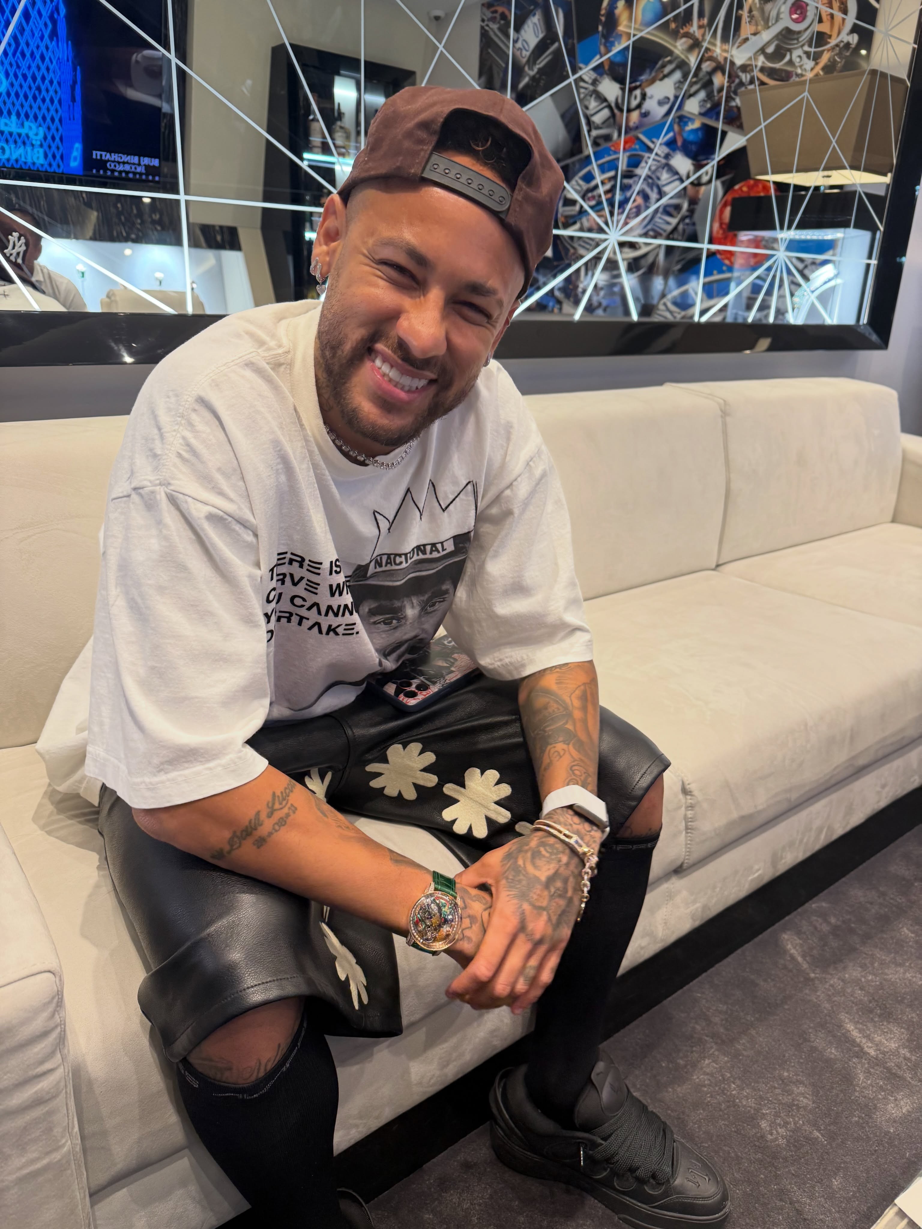 Neymar Jr. sorrindo, sentado em um sofá bege, usando boné marrom, camiseta branca estampada, bermuda de couro preta com detalhes de flores brancas, meias e tênis pretos. Ele exibe tatuagens nos braços e pulsos, além de um relógio chamativo