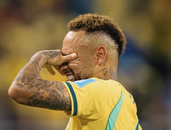 Jogador de futebol com cabelo crespo e loiro, suado, cobrindo o rosto com a mão tatuada, expressão de angústia, vestindo camisa amarela com detalhes azuis, em campo