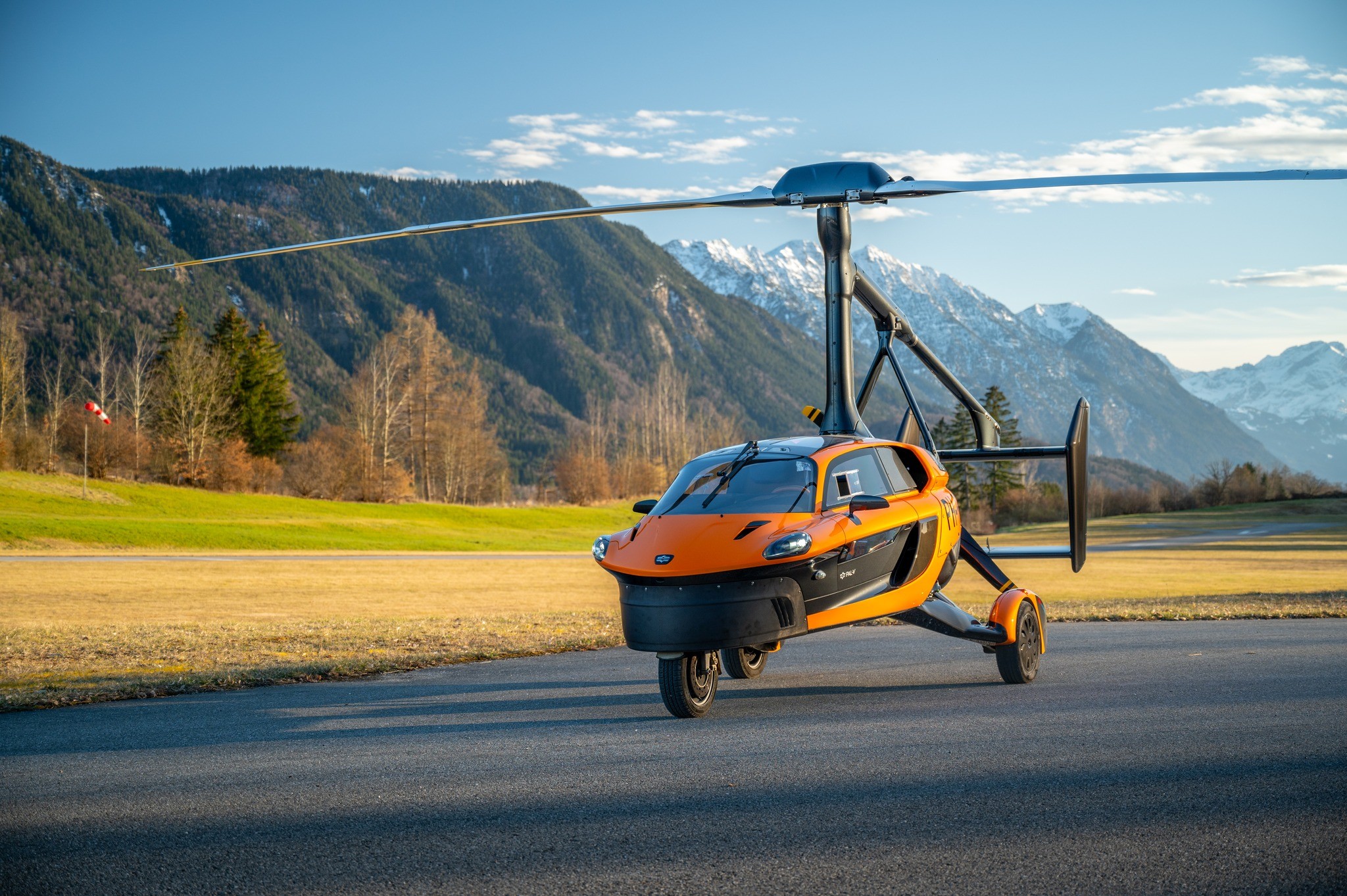 Um carro voador laranja e preto, com hélices de helicóptero, parado em uma pista de asfalto. Ao fundo, montanhas verdes e nevadas sob um céu azul claro. A paisagem inclui árvores e grama seca, com uma bandeira vermelha à esquerda. O veículo tem três rodas e design futurista.