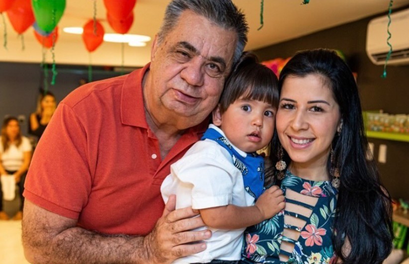 Homem idoso de camisa polo vermelha abraça um bebê de camisa branca e lenço azul, que está no colo de uma mulher sorridente com vestido floral e cabelo escuro, em um ambiente decorado com balões vermelhos e verdes