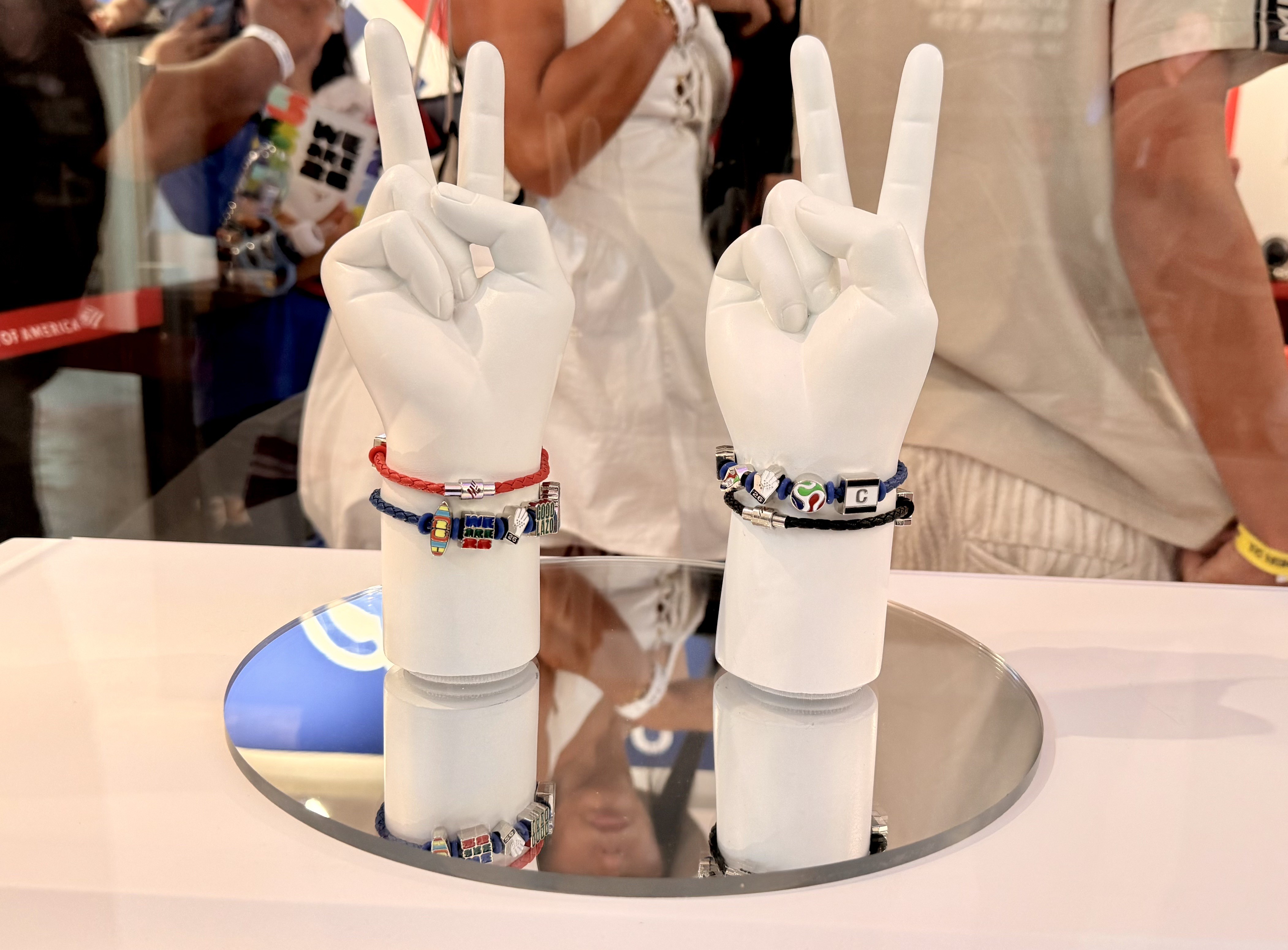 Duas mãos de manequim brancas, em formato de V de vitória, exibem pulseiras coloridas com pingentes temáticos sobre uma superfície espelhada. A mão esquerda tem uma pulseira vermelha e outra azul com pingentes prateados e coloridos. A mão direita exibe uma pulseira preta e outra azul com pingentes de esferas e um quadrado preto com a letra C. Pessoas desfocadas aparecem ao fundo