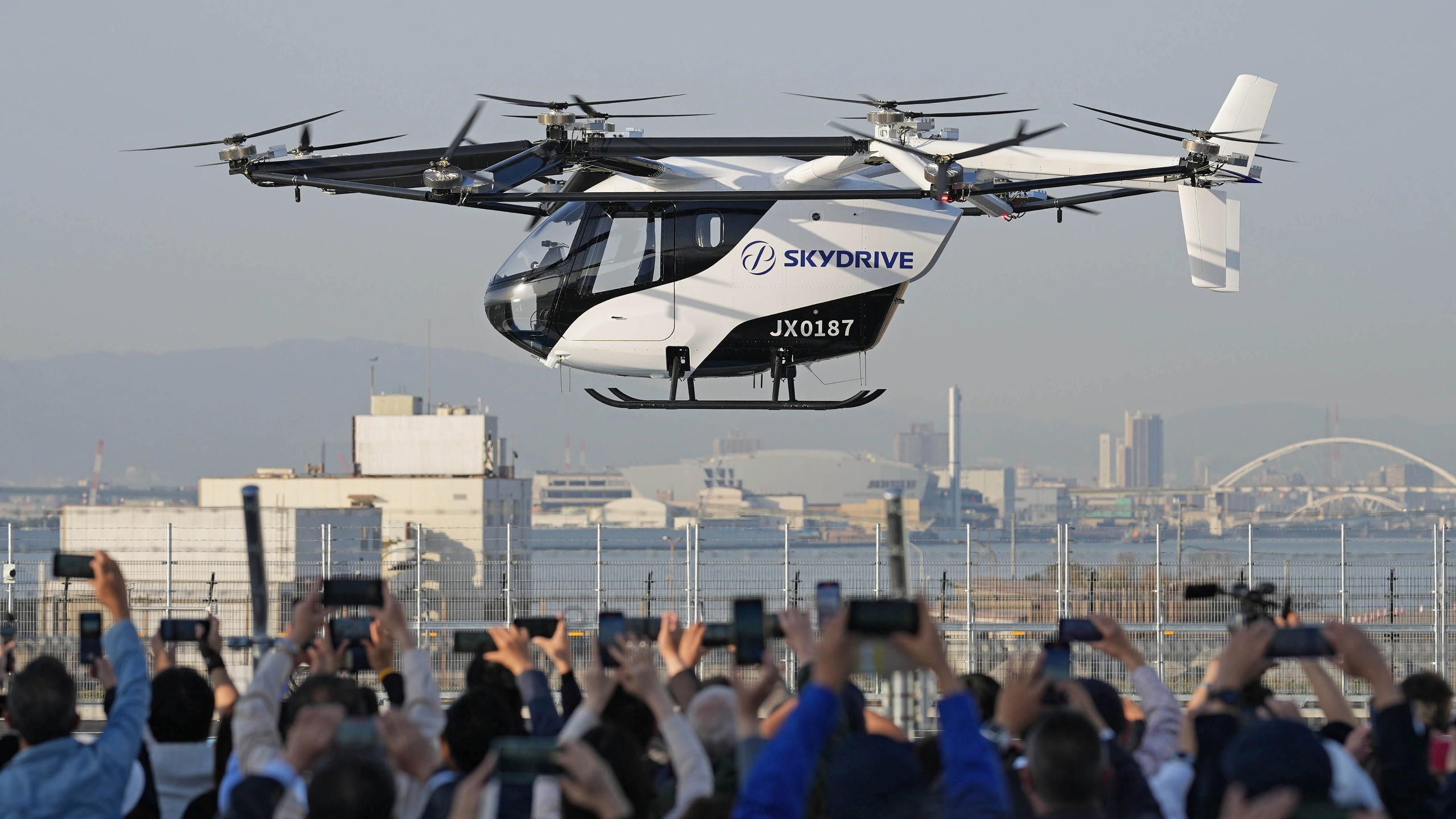 Um protótipo de carro voador branco e preto da Skydrive, modelo JX0187, paira no ar sobre uma multidão que o fotografa com celulares. Ao fundo, uma cidade com prédios e uma ponte arqueada sob um céu claro