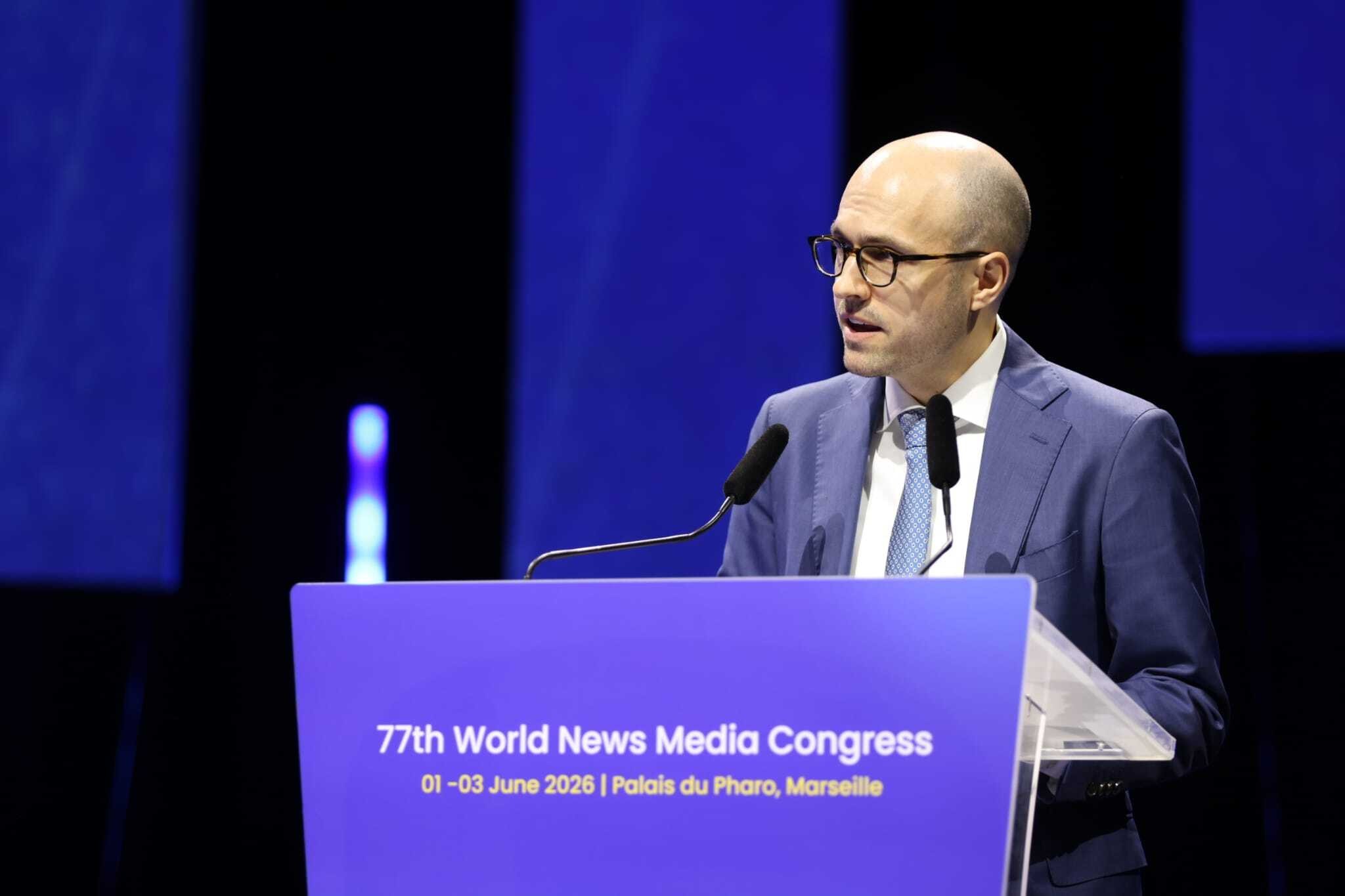 Homem calvo de óculos e terno azul, gravata clara, discursando em púlpito azul com "77th World News Media Congress" e data