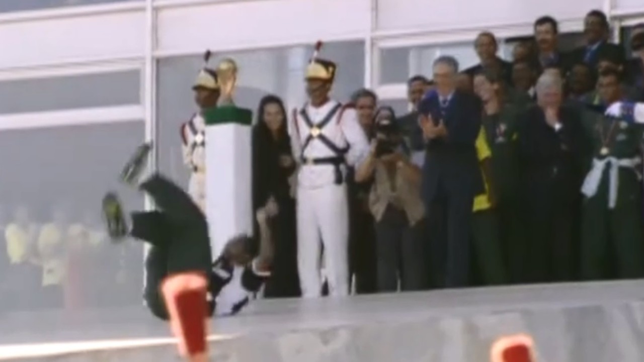 Um homem de camisa branca e calça escura caindo de costas, com as pernas para cima, em frente a uma multidão de pessoas em um evento formal. Ao fundo, um militar de uniforme branco e chapéu dourado observa a cena, com outros convidados e seguranças ao redor