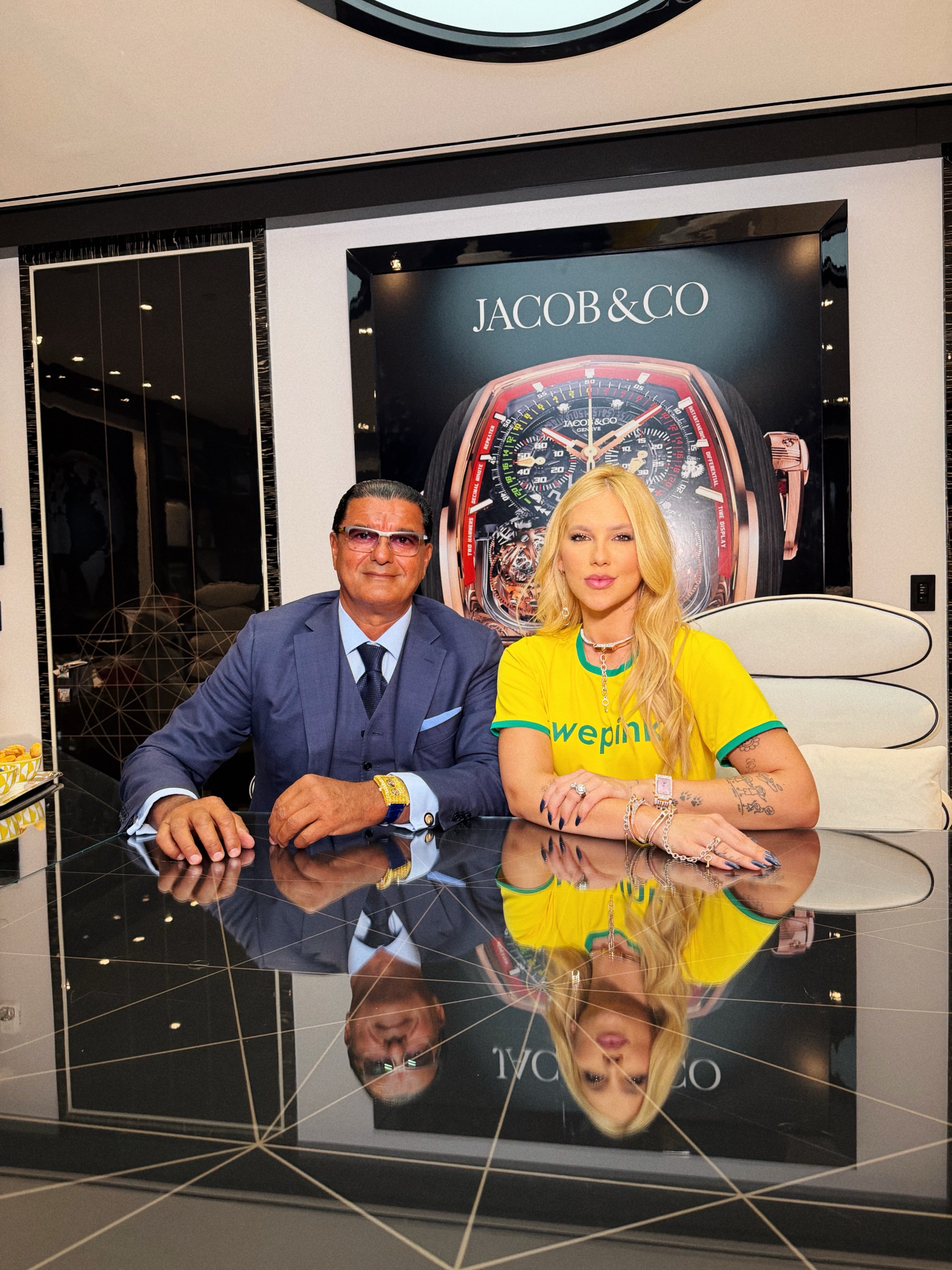 Um homem e uma mulher posam sentados à mesa preta espelhada. Ele usa terno azul, óculos e relógio dourado. Ela veste camiseta amarela com wepink e joias. Ao fundo, um pôster de relógio Jacob & Co