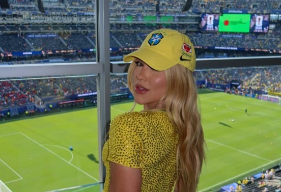 Mulher loira de boné amarelo do Brasil e blusa amarela brilhante, olhando para trás com um sorriso, em um camarote de estádio de futebol lotado
