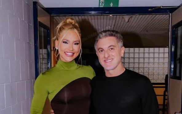 Apresentadora loira de coque alto, maquiagem marcante e blusa verde e marrom, sorri ao lado de apresentador de cabelo grisalho e camiseta preta, ambos em frente a uma porta com sinalização de saída de emergência