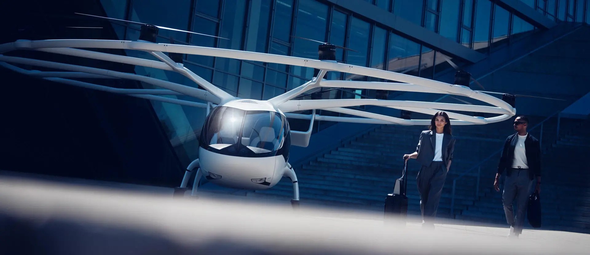 Um eVTOL branco, com múltiplos rotores, estacionado em frente a um prédio moderno de vidro azul. À direita, uma mulher de terno cinza puxa uma mala de rodinhas, e um homem de jaqueta escura e camiseta branca caminha ao lado, carregando uma pasta.