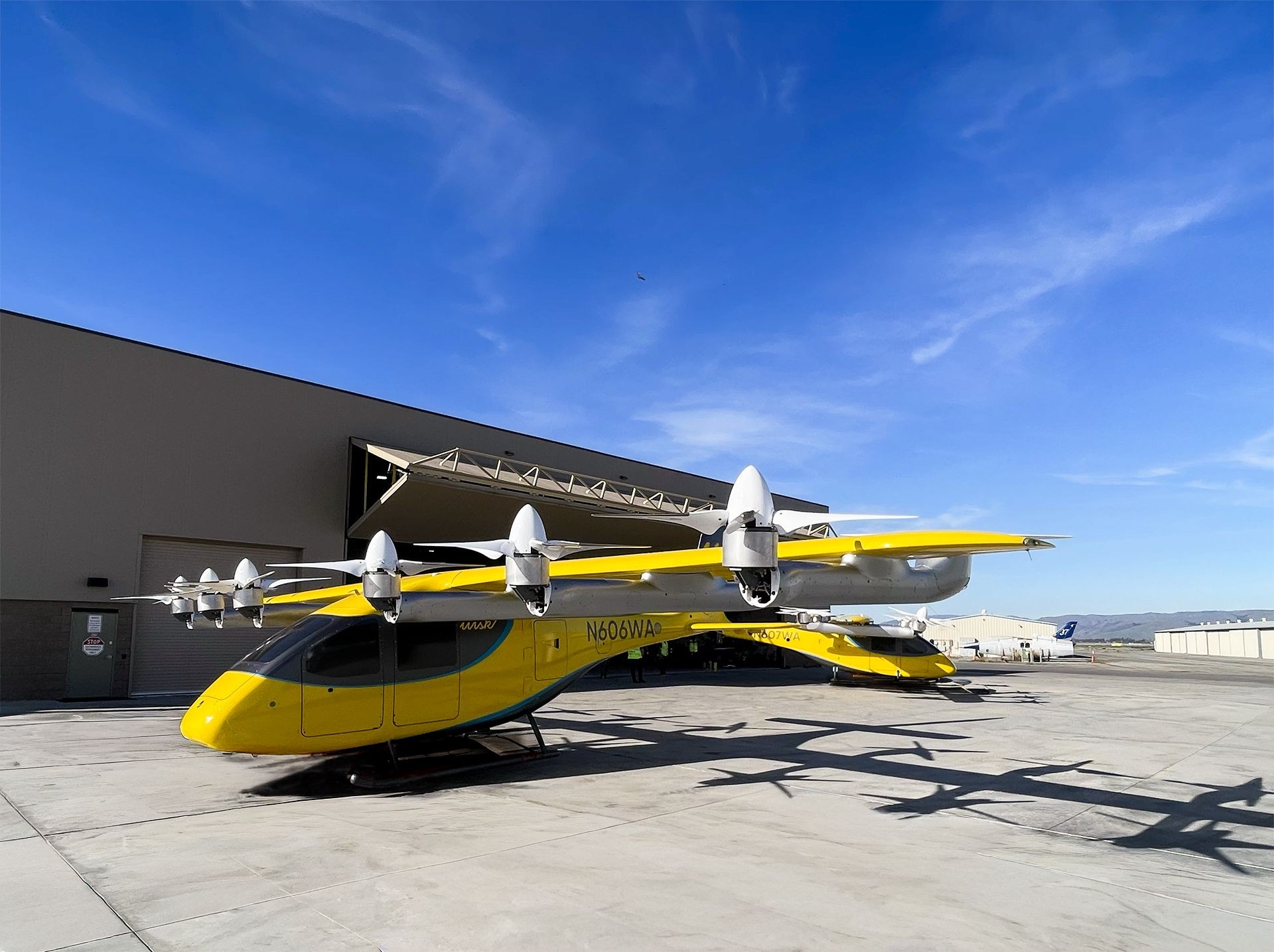 Aeronave elétrica amarela e cinza com oito hélices em suas asas, estacionada em uma pista de concreto, com um hangar ao fundo sob céu azul claro