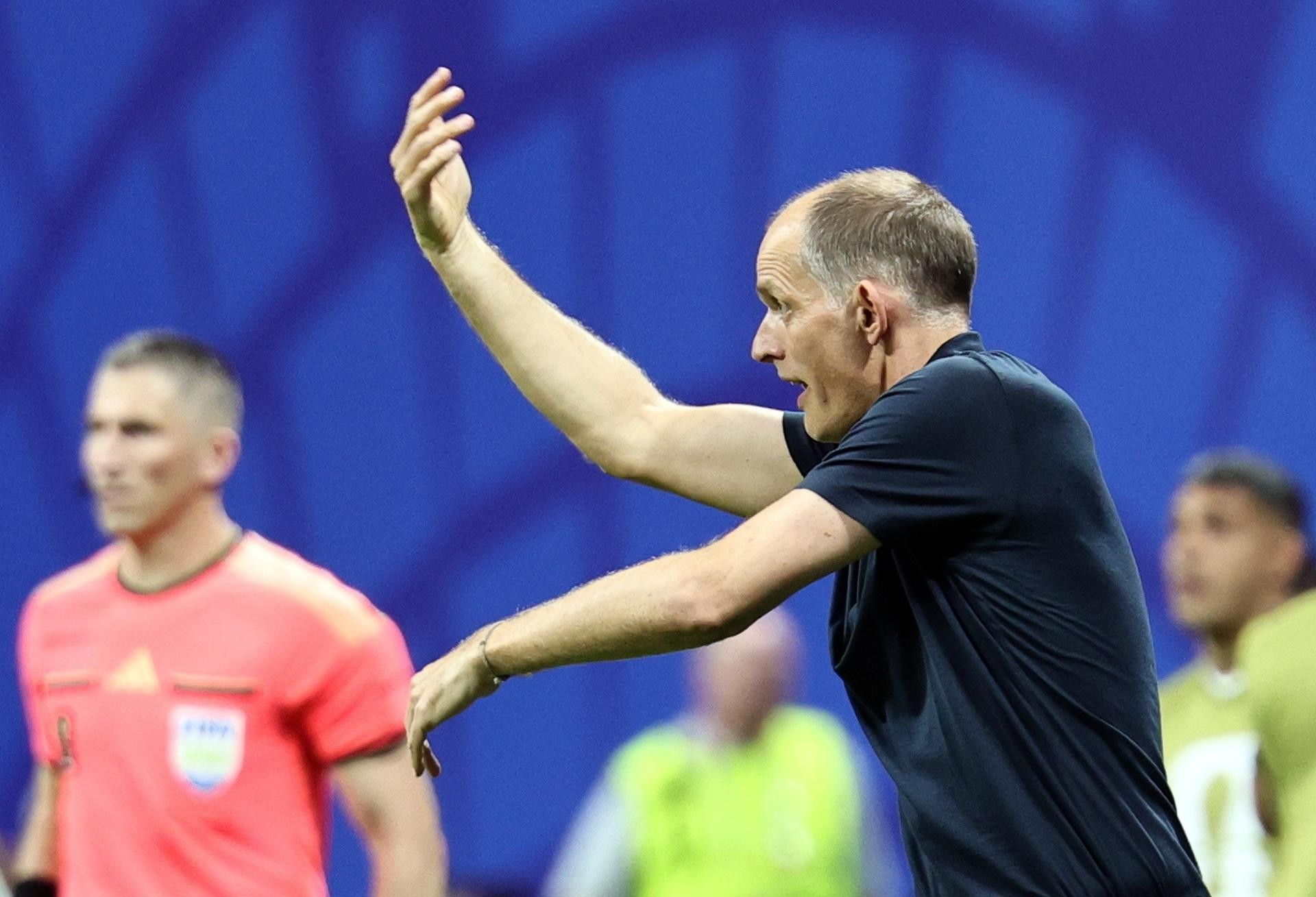 Homem careca de camisa azul escura, com a boca aberta e braços erguidos, gesticulando intensamente em campo de futebol azul. Ao fundo, um árbitro de camisa laranja e outros jogadores desfocados