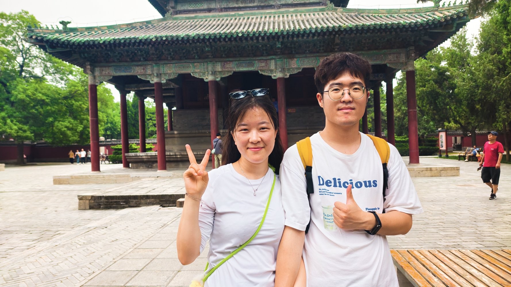 Um casal asiático sorridente posa em frente a um templo chinês. A mulher, à esquerda, veste camiseta branca e faz sinal de paz. O homem, à direita, usa óculos, camiseta branca com a palavra Delicious e faz sinal de positivo. Ambos têm mochilas e óculos de sol na cabeça da mulher. O templo tem telhado verde e pilares vermelhos, com árvores ao fundo