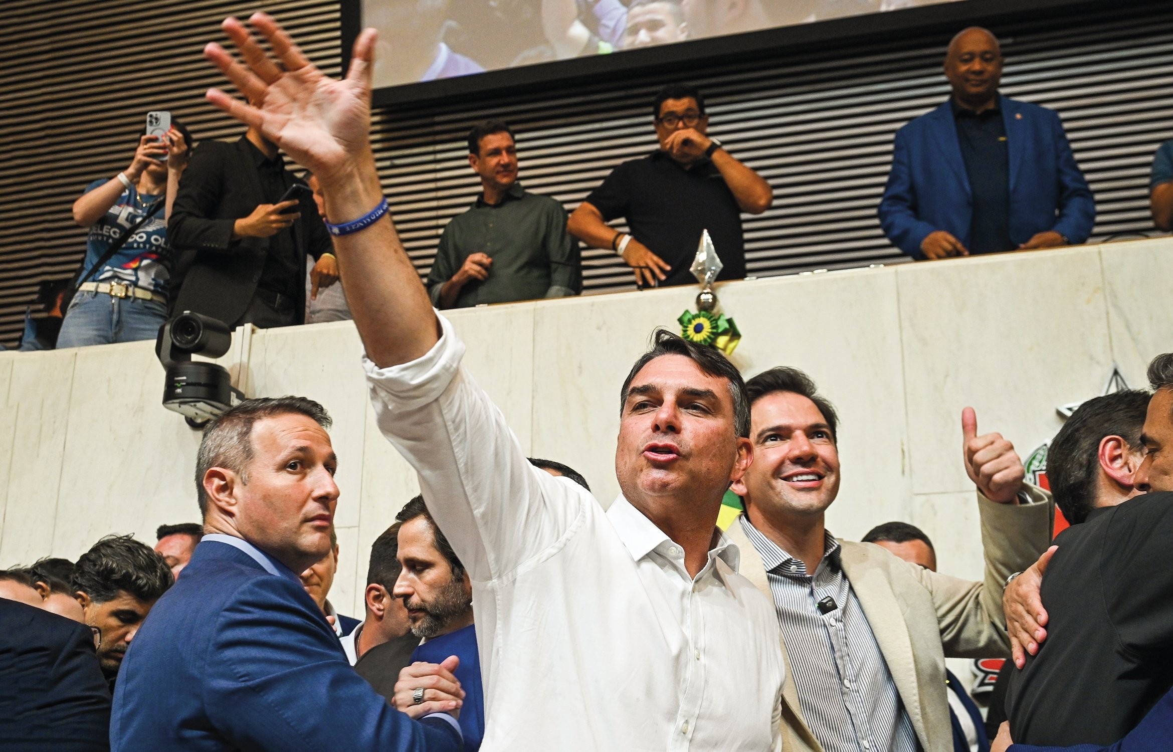 Jair Bolsonaro, de camisa branca, acena com a mão direita levantada, em meio a uma multidão. Ao seu lado, um homem de blazer bege sorri e faz um sinal de positivo. Outros homens observam de um balcão superior, incluindo um de blazer azul. O ambiente parece ser um evento político ou público, com pessoas filmando e fotografando.