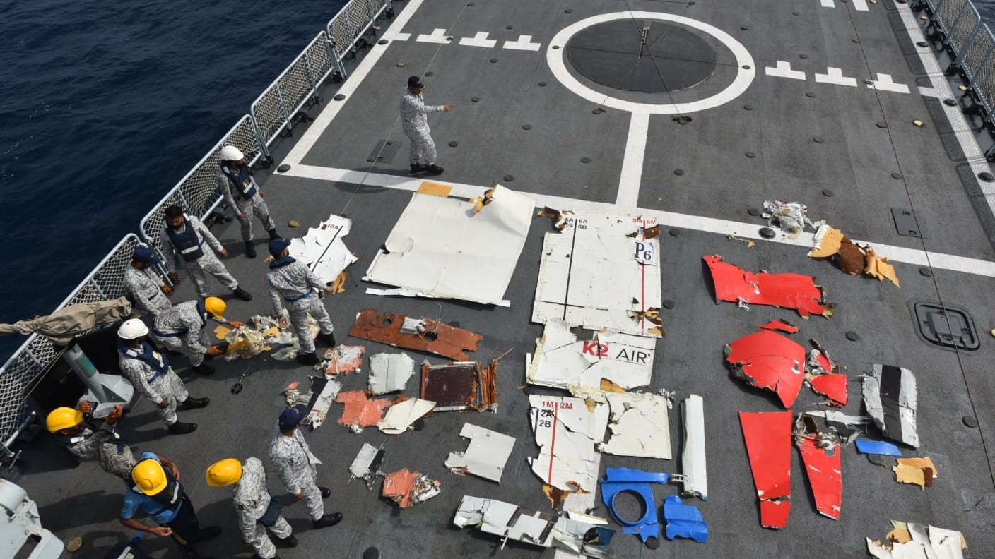 Militares em uniformes camuflados e coletes de seguran&ccedil;a inspecionam destro&ccedil;os de avi&atilde;o, incluindo pe&ccedil;as brancas, vermelhas e azuis, espalhados no conv&eacute;s cinza de um navio, com o oceano azul ao lado