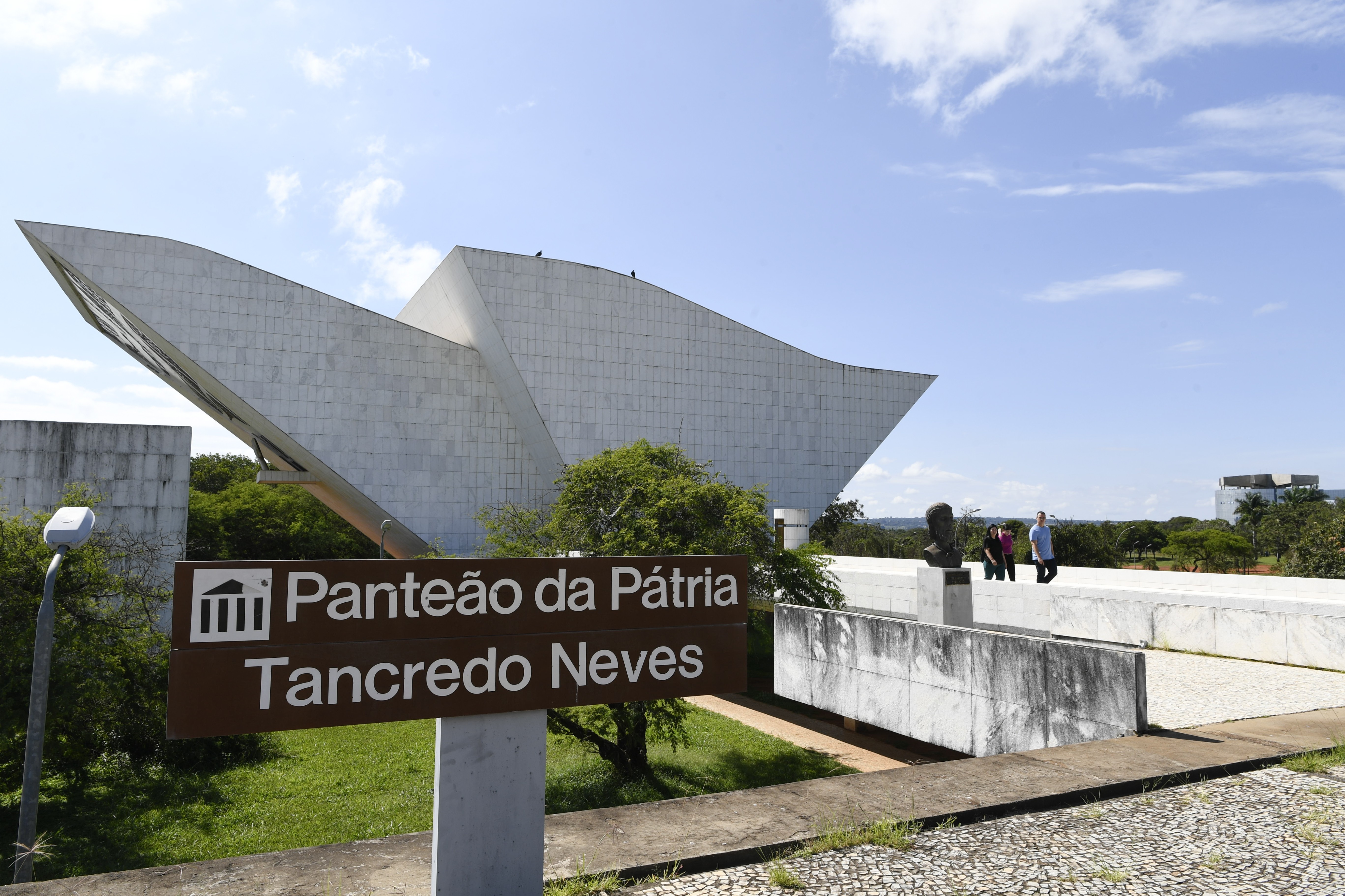 Placa marrom com Panteão da Pátria Tancredo Neves em branco, em frente ao edifício branco de arquitetura moderna, com formas angulares. O céu azul claro tem nuvens esparsas, e há árvores verdes ao redor. Três pessoas e um busto escuro são visíveis na plataforma do panteão.