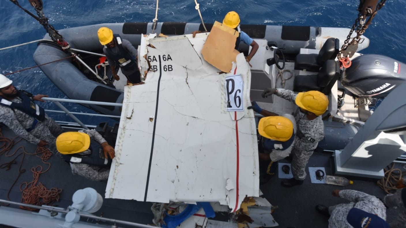 Membros da equipe de resgate, usando capacetes de seguran&ccedil;a e coletes salva-vidas, manuseiam um grande peda&ccedil;o branco de destro&ccedil;o de avi&atilde;o com a inscri&ccedil;&atilde;o P6 e 5A 6A 5B 6B em um barco no mar