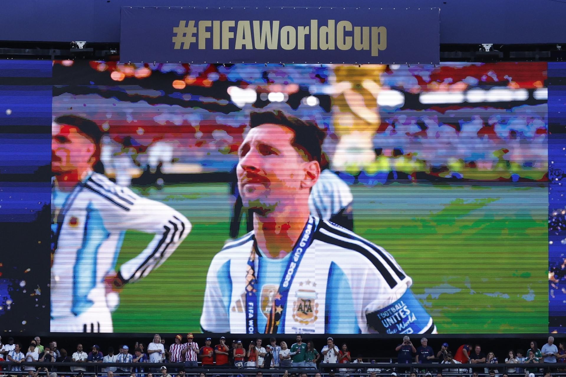 Lionel Messi, jogador argentino, em close-up na tela de um telão, usando camisa branca e azul da seleção, com medalha no pescoço. Ele olha para cima, com expressão séria. Ao fundo, um campo de futebol e torcedores. Acima, uma faixa azul com a hashtag #FIFAWorldCup. Abaixo, uma arquibancada com público