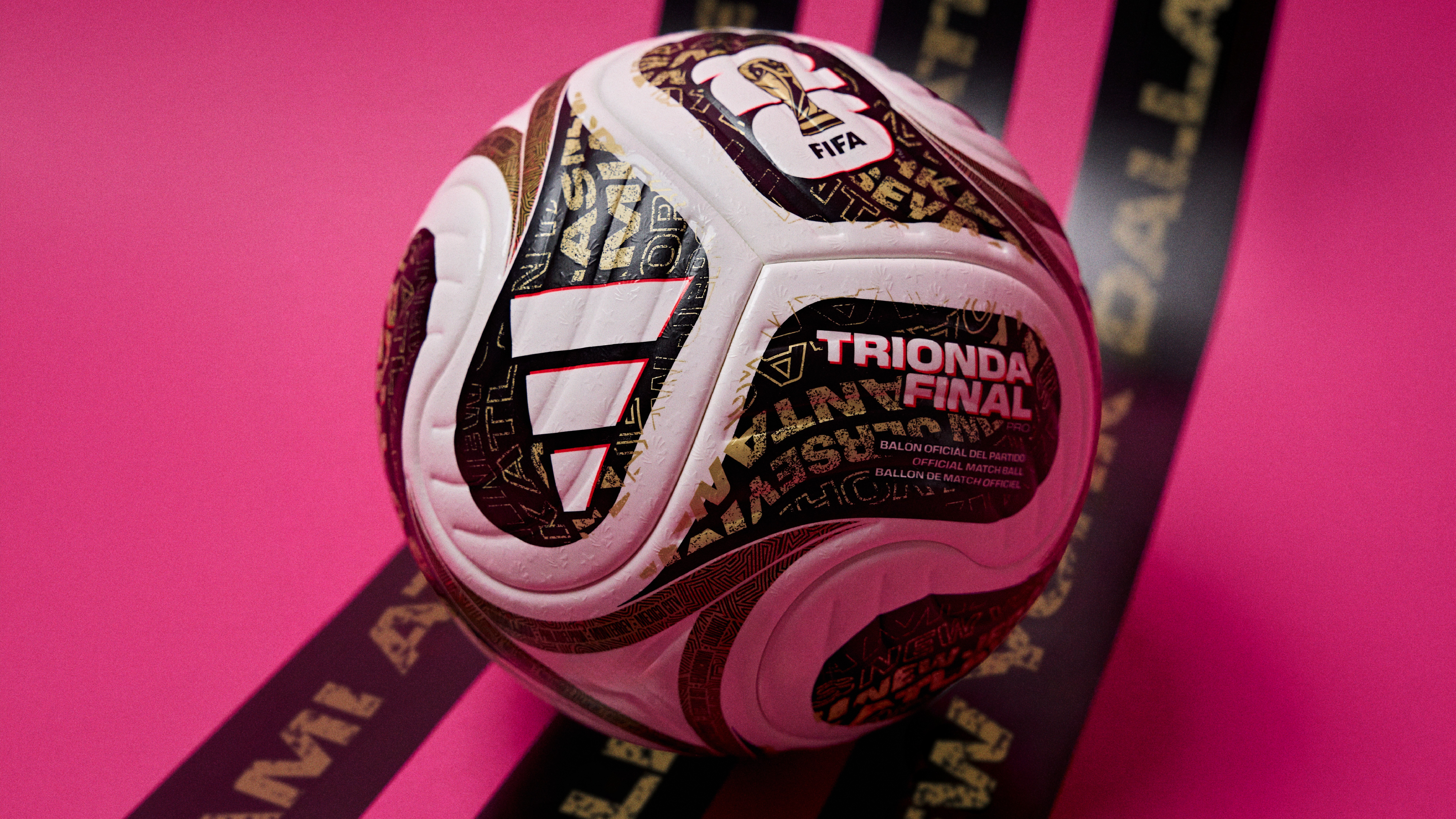 Bola de futebol Adidas Brazuca Final Rio, branca com detalhes dourados e pretos, sobre fundo rosa vibrante e duas faixas pretas com letras douradas