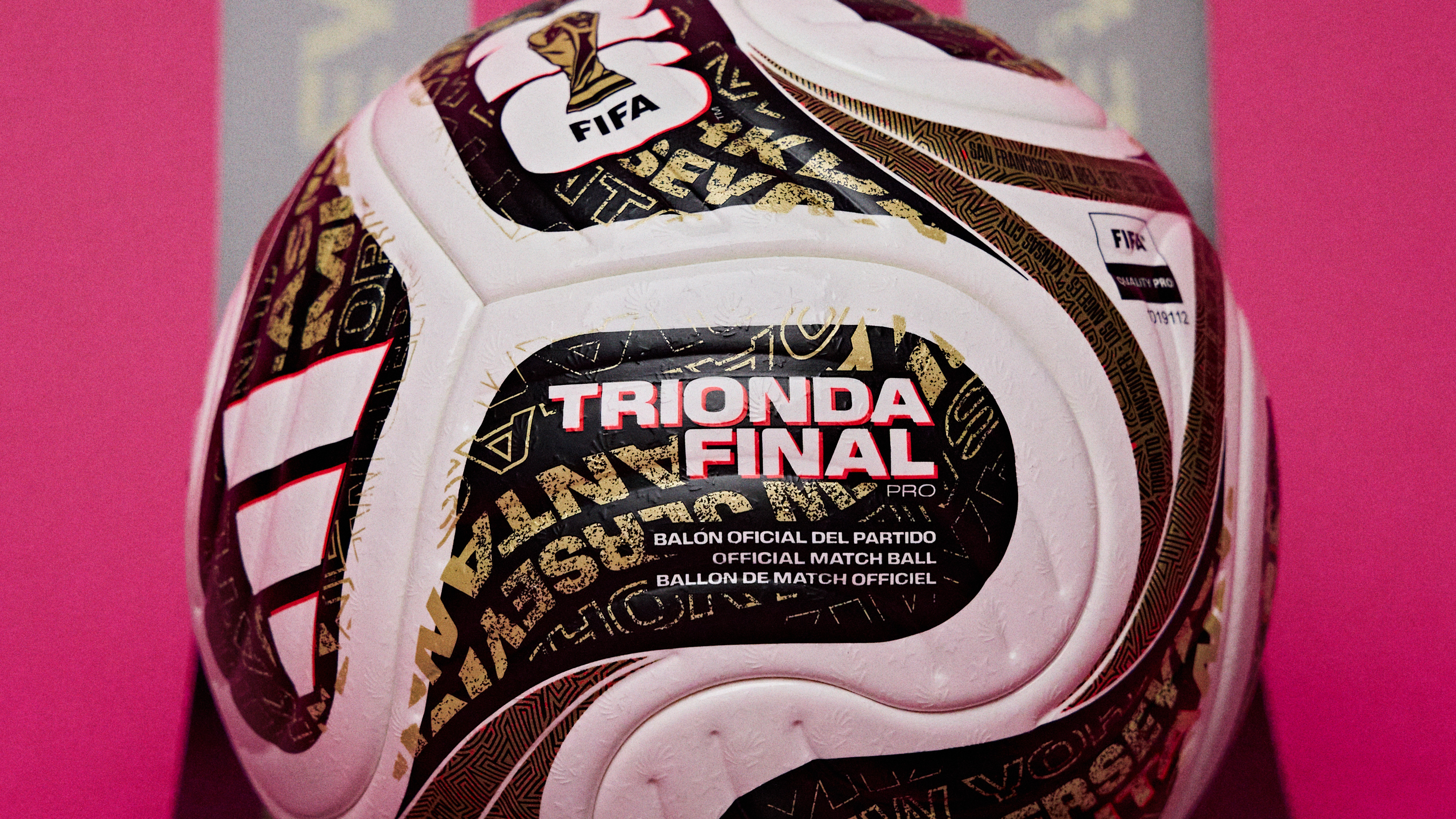 Bola de futebol branca com detalhes em preto e dourado, incluindo o logo da FIFA e o texto TRIO NDA FINAL PRO em destaque, sobre um fundo rosa