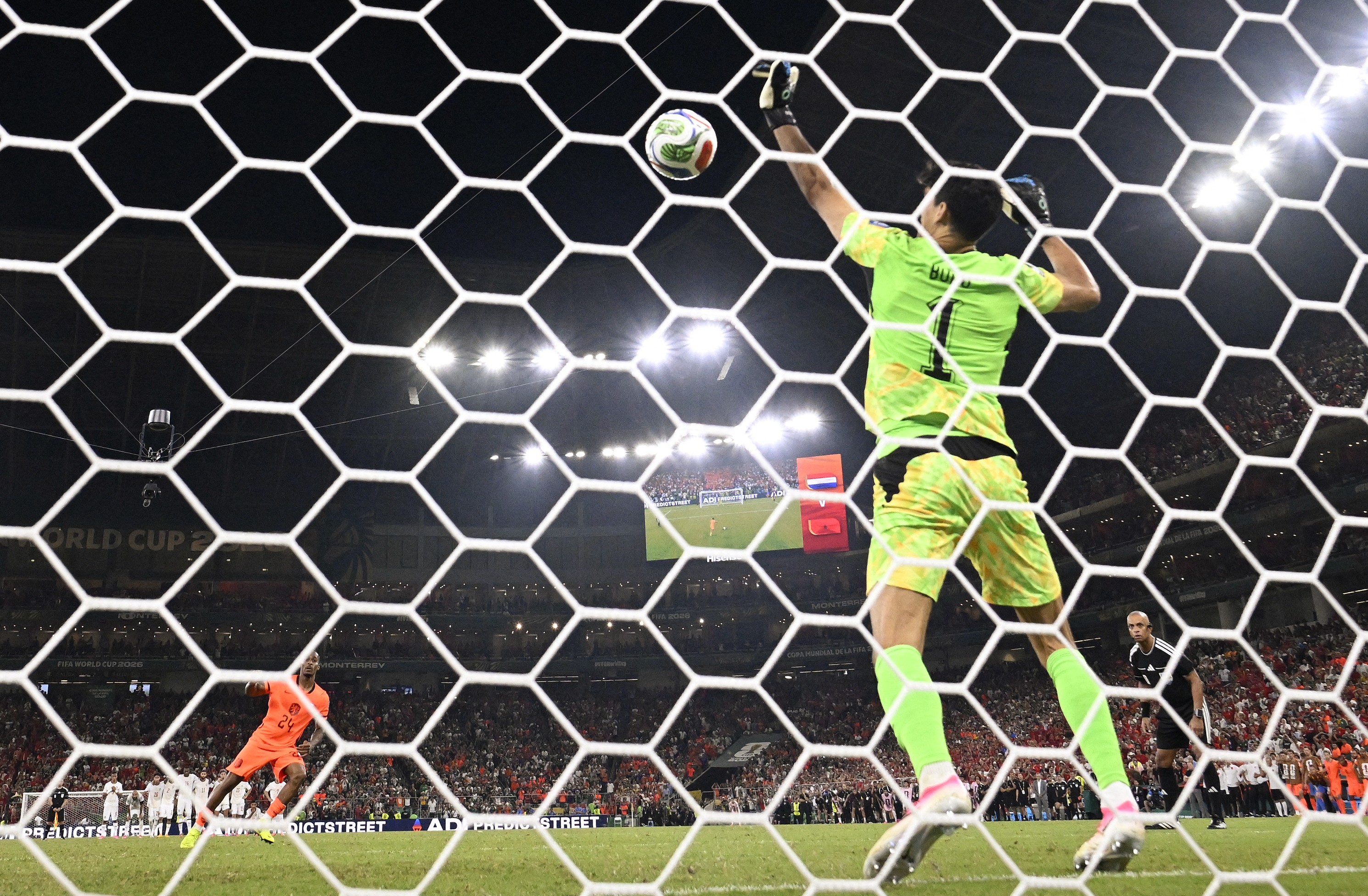 Goleiro de camisa verde neon salta para defender uma bola de futebol no alto, vista através da rede hexagonal branca do gol, em um estádio lotado e iluminado à noite