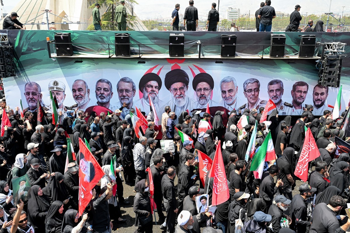 Multidão de pessoas, muitas vestindo preto, segurando bandeiras iranianas e vermelhas com inscrições árabes, em frente a um grande banner com retratos de líderes iranianos. Alguns homens estão em uma plataforma acima da multidão