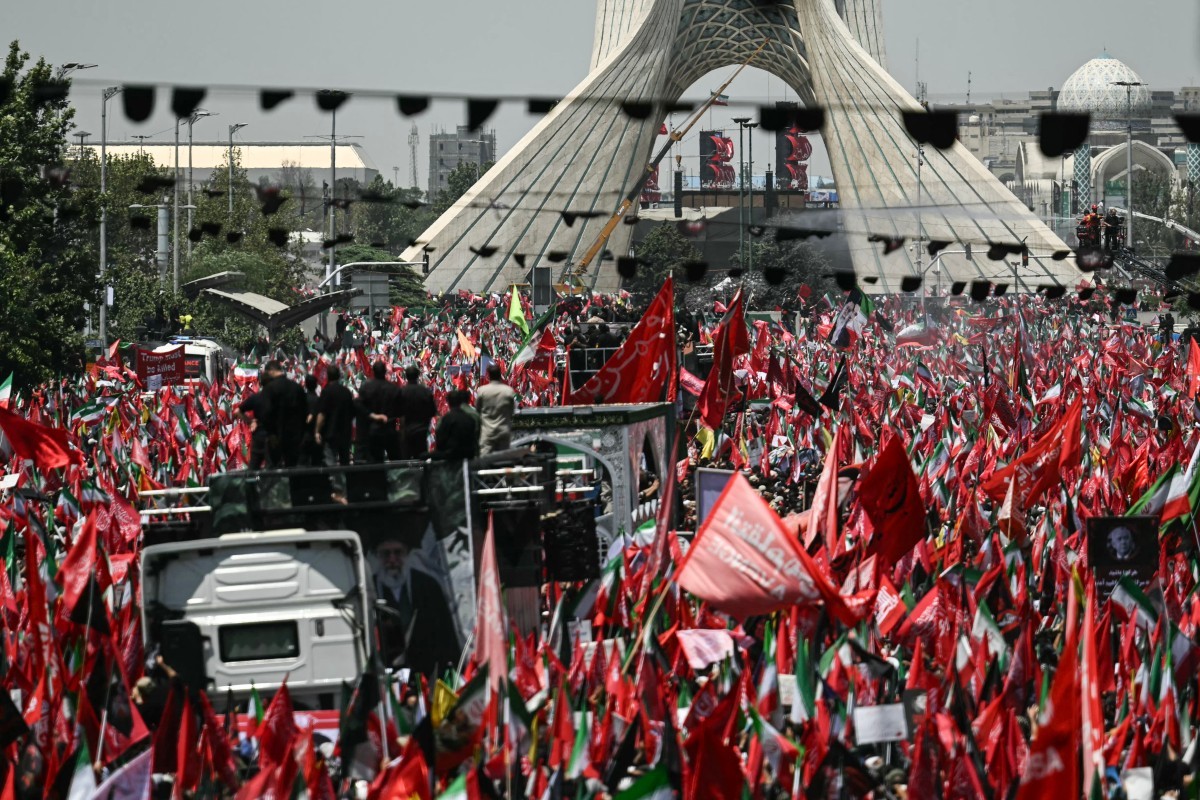 Multidão de pessoas em protesto, com milhares de bandeiras vermelhas e algumas verdes, brancas e pretas, em frente à Torre Azadi em Teerã. Homens em cima de um caminhão branco observam a manifestação. O céu está claro e o ambiente é de grande agitação