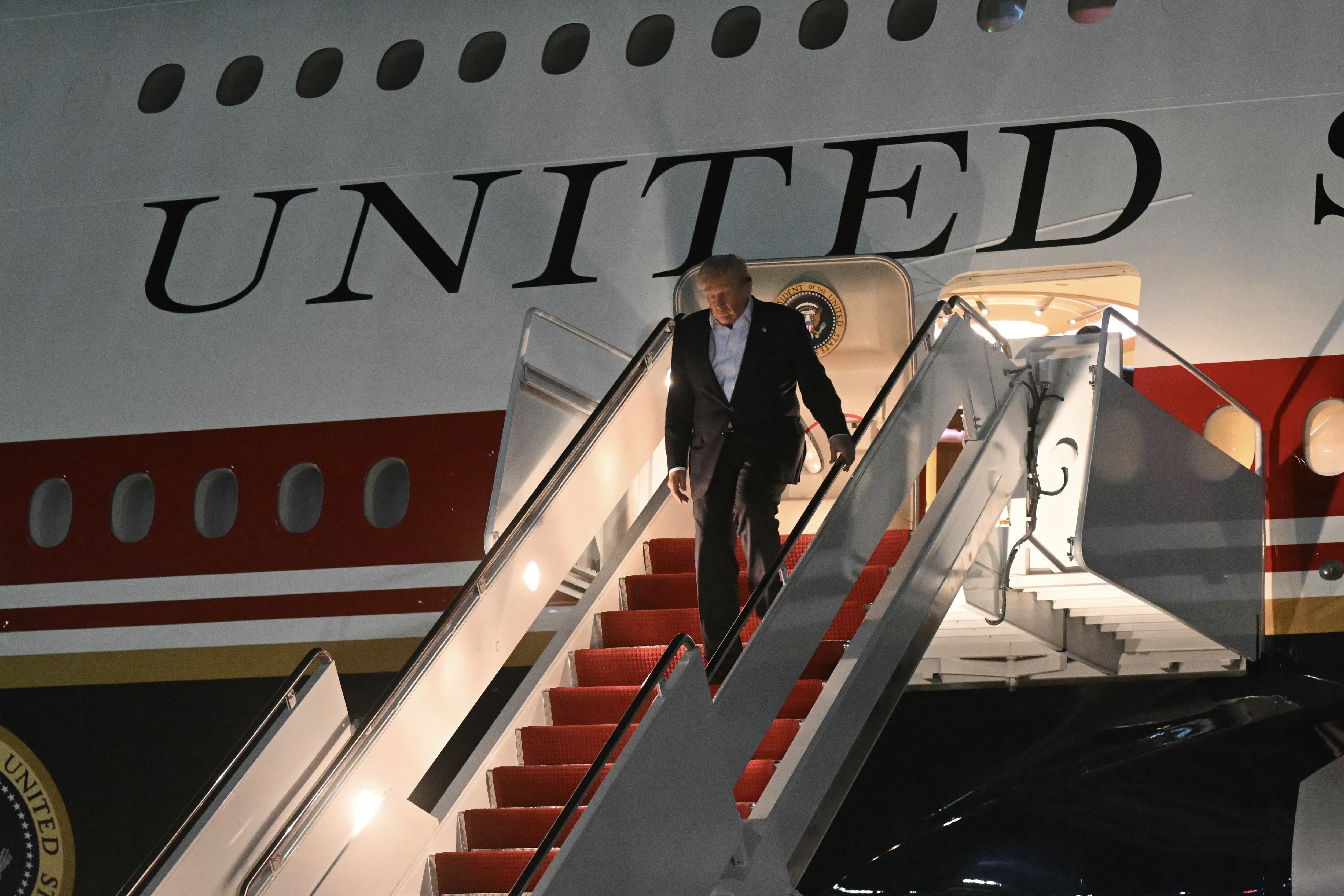 Homem de terno escuro e camisa clara descendo a escada vermelha de um avião branco com a palavra UNITED em letras pretas na lateral