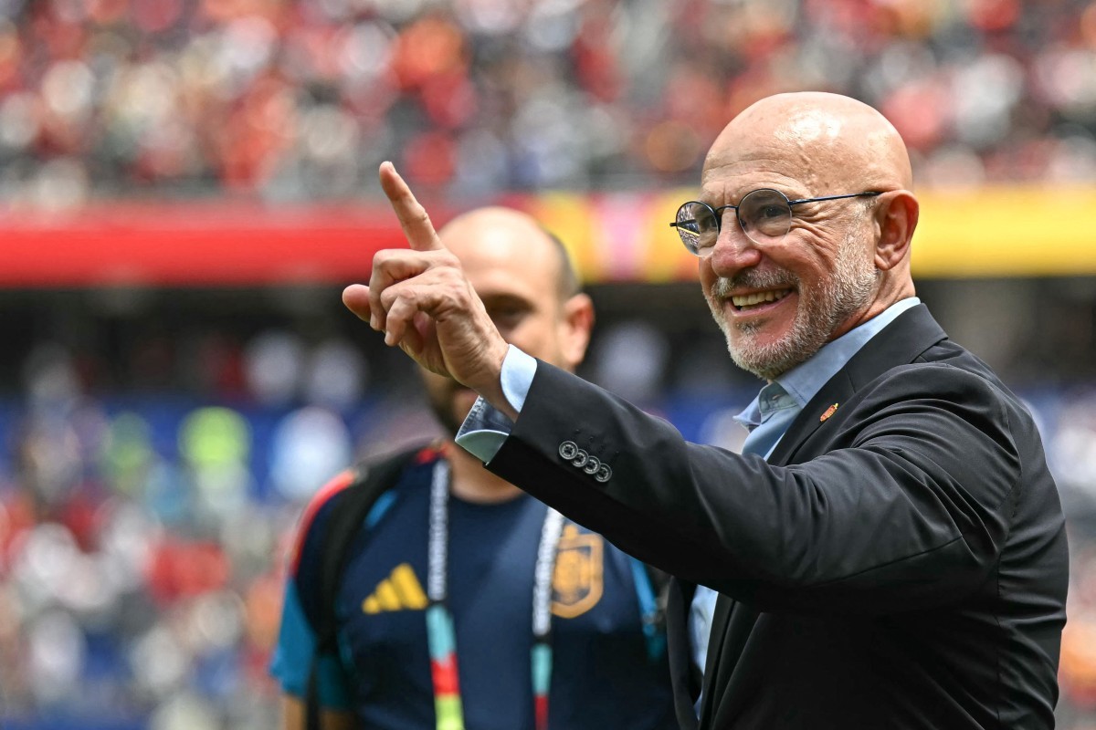 Homem careca de óculos e barba grisalha, sorrindo e apontando para cima com o indicador direito, vestindo terno preto e camisa azul clara, em um estádio com público desfocado ao fundo