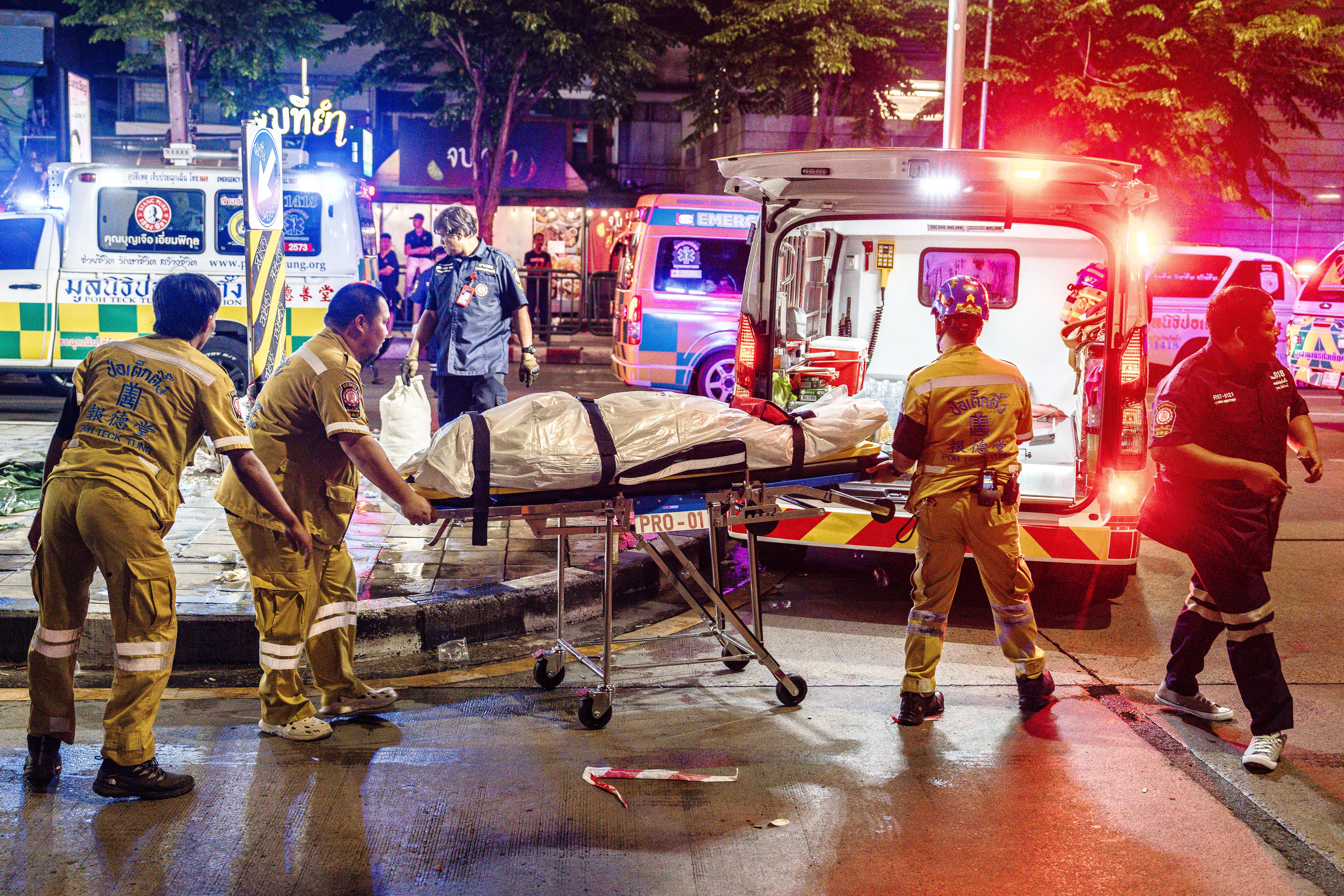 Equipe de resgate tailandesa, com uniformes amarelos e azuis, movimenta uma maca com um corpo coberto em uma rua molhada, com ambulâncias e luzes de emergência ao fundo, à noite