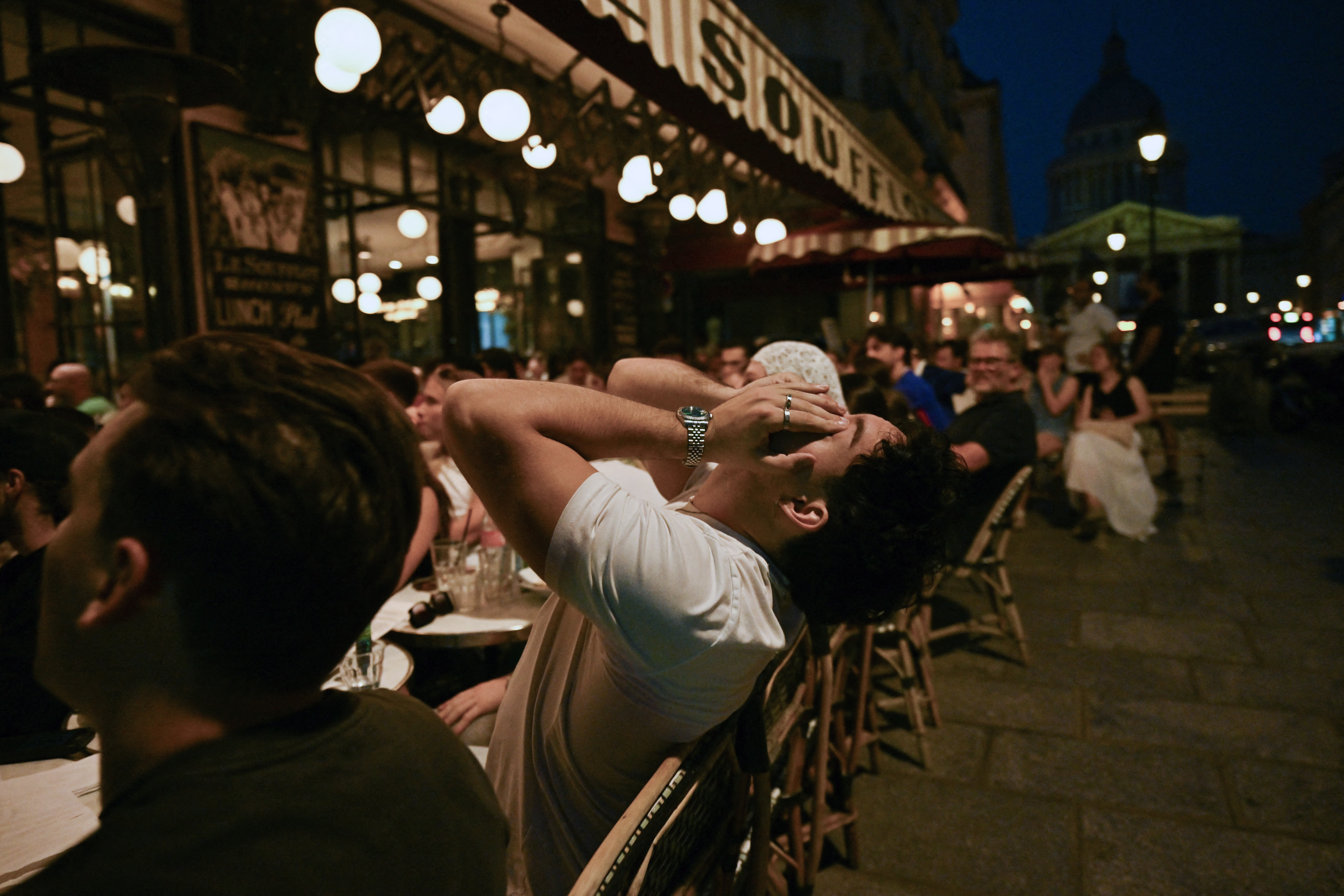 Homem de camiseta branca, cabelo cacheado, com as mãos na cabeça e olhos fechados, sentado em uma cadeira de madeira em uma calçada de café parisiense à noite. Ao fundo, pessoas sentadas em mesas iluminadas e um edifício com cúpula