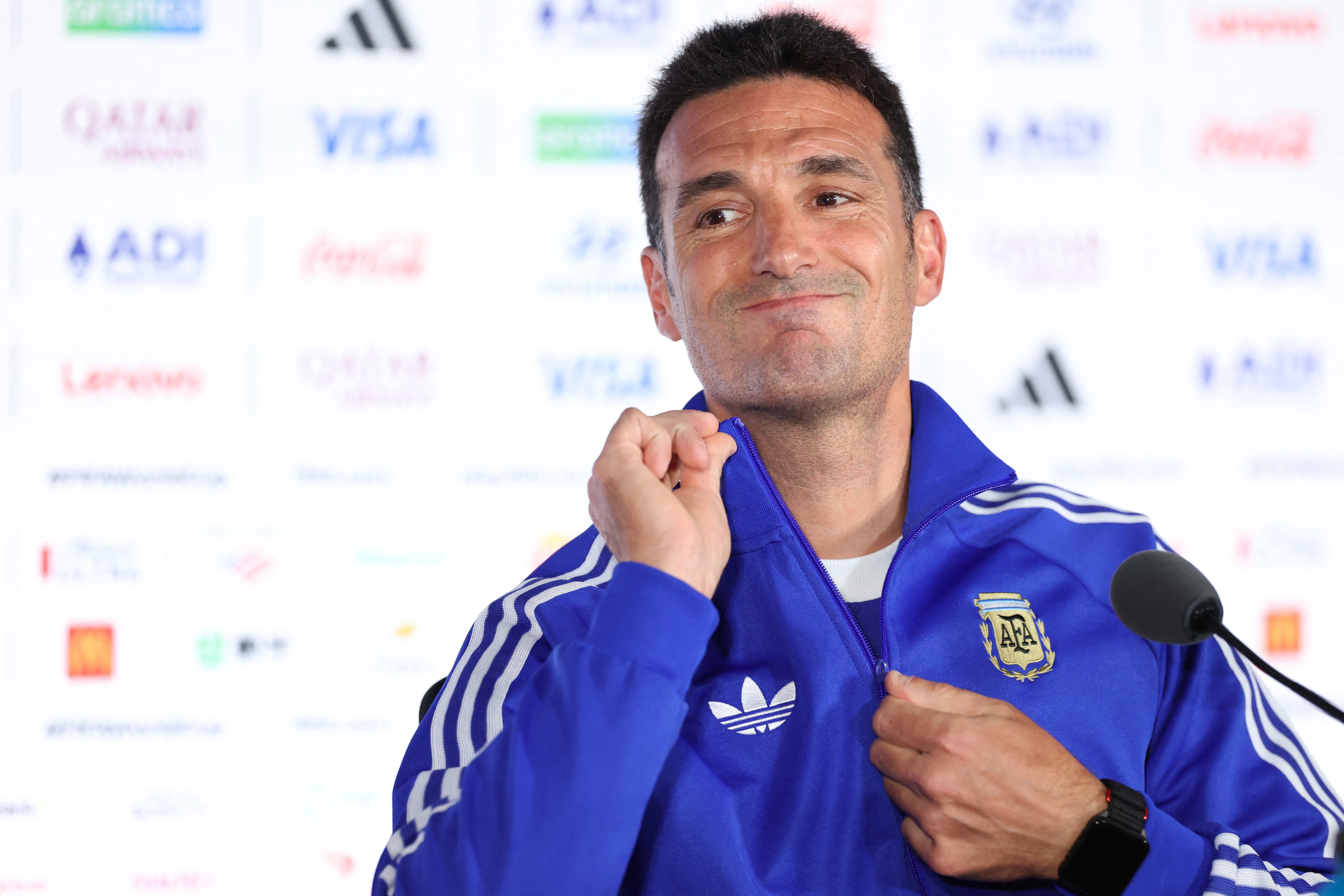 Homem de pele clara, cabelo escuro e barba rala, vestindo agasalho azul com listras brancas e o escudo da AFA, sorri levemente enquanto ajusta o zíper. Ele está em frente a um painel branco com logotipos de patrocinadores como Qatar Airways, Visa e Adidas, e um microfone preto aparece no canto direito