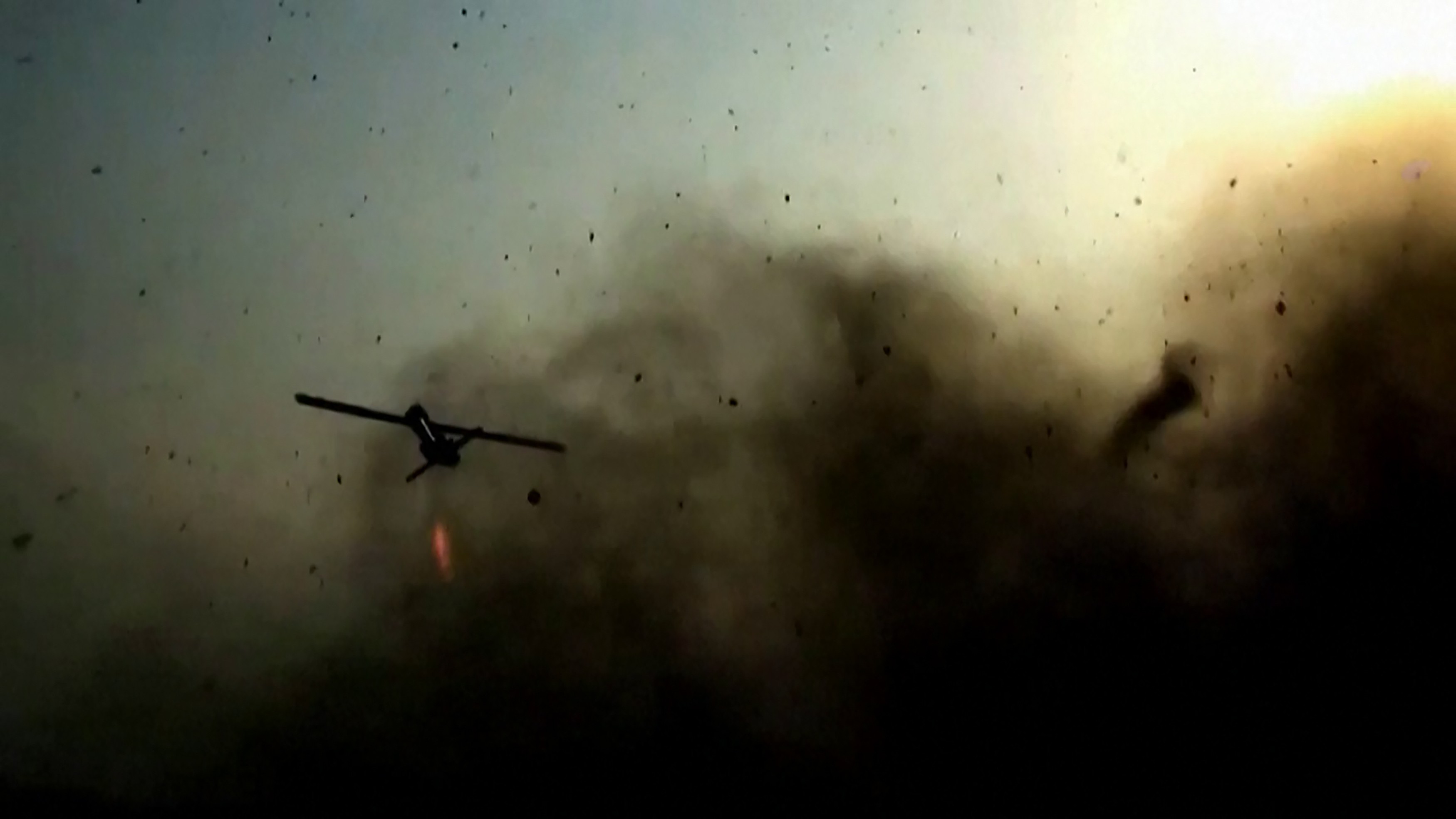 Um drone militar escuro, com asas finas e uma luz vermelha embaixo, voa em meio a uma nuvem de fumaça e detritos. O céu claro e amarelado no canto superior direito contrasta com a escuridão da fumaça, que domina a parte inferior e central da imagem, sugerindo um cenário de conflito ou destruição