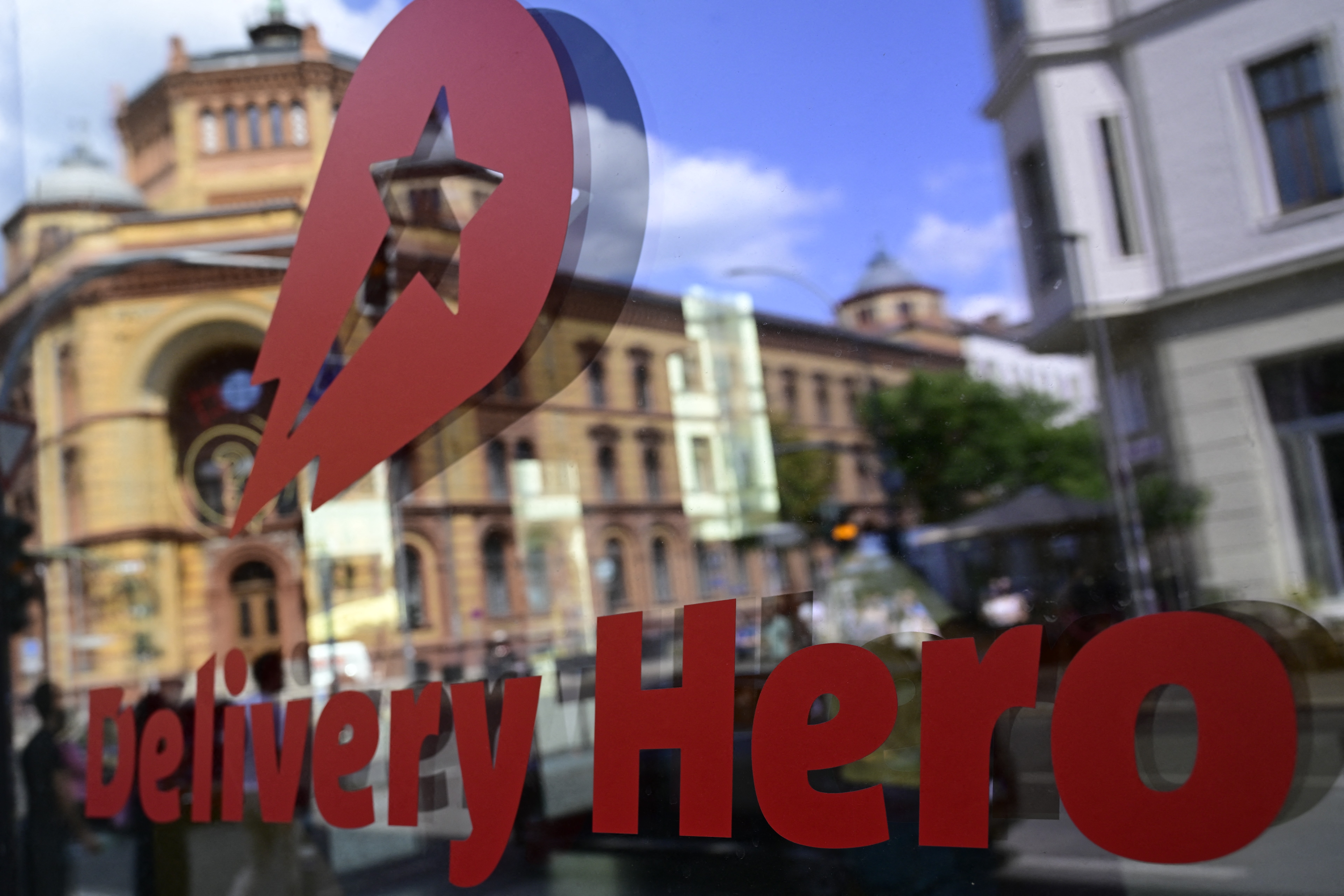 O logotipo vermelho da Delivery Hero e um ícone de estrela com um raio são refletidos em uma janela, com prédios antigos e o céu azul ao fundo