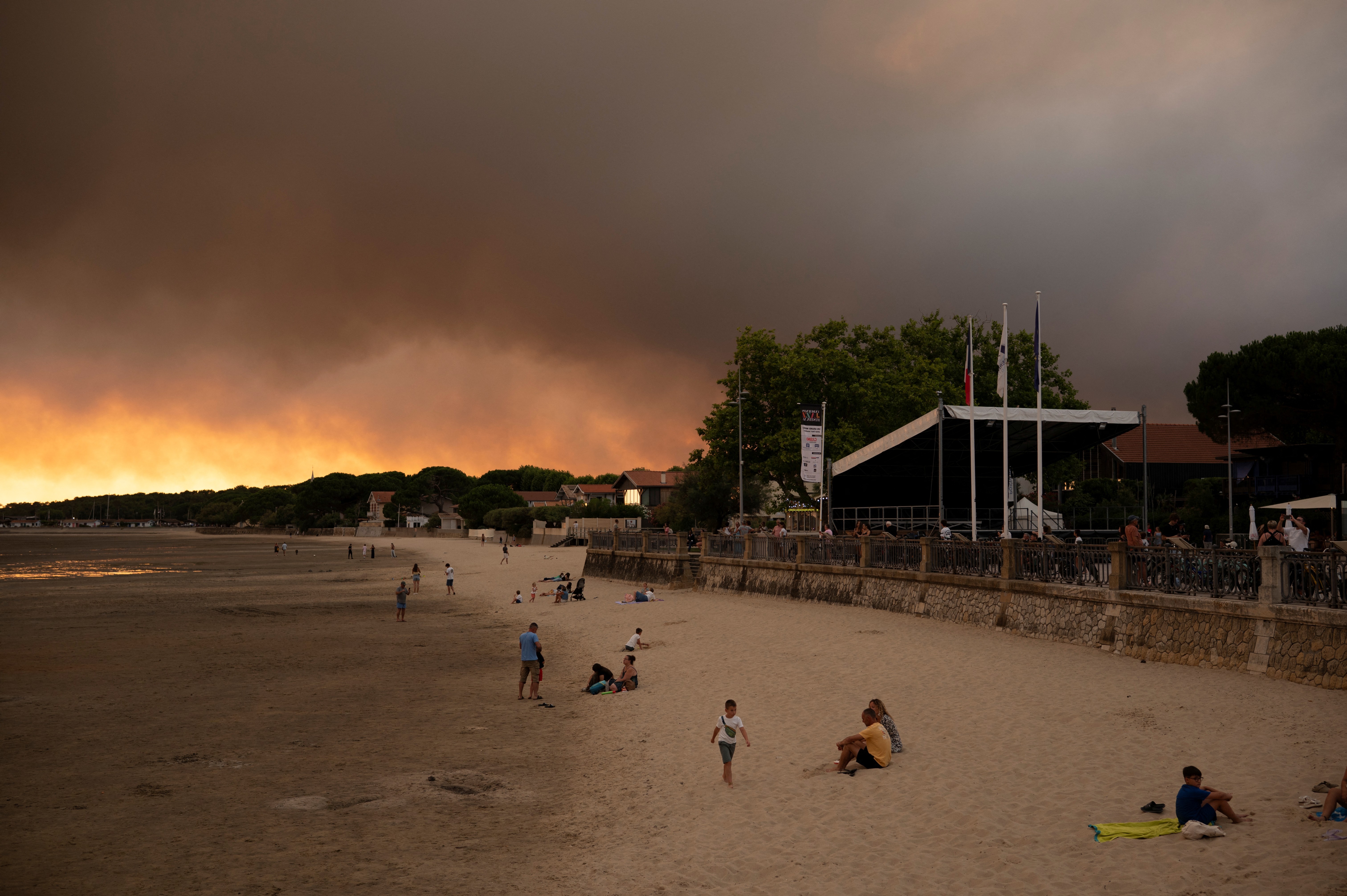 Céu escuro e alaranjado sobre uma praia com areia clara, onde pessoas relaxam e caminham. À direita, um calçadão com um prédio de telhado escuro e bandeiras hasteadas. Ao fundo, uma faixa de árvores e casas.