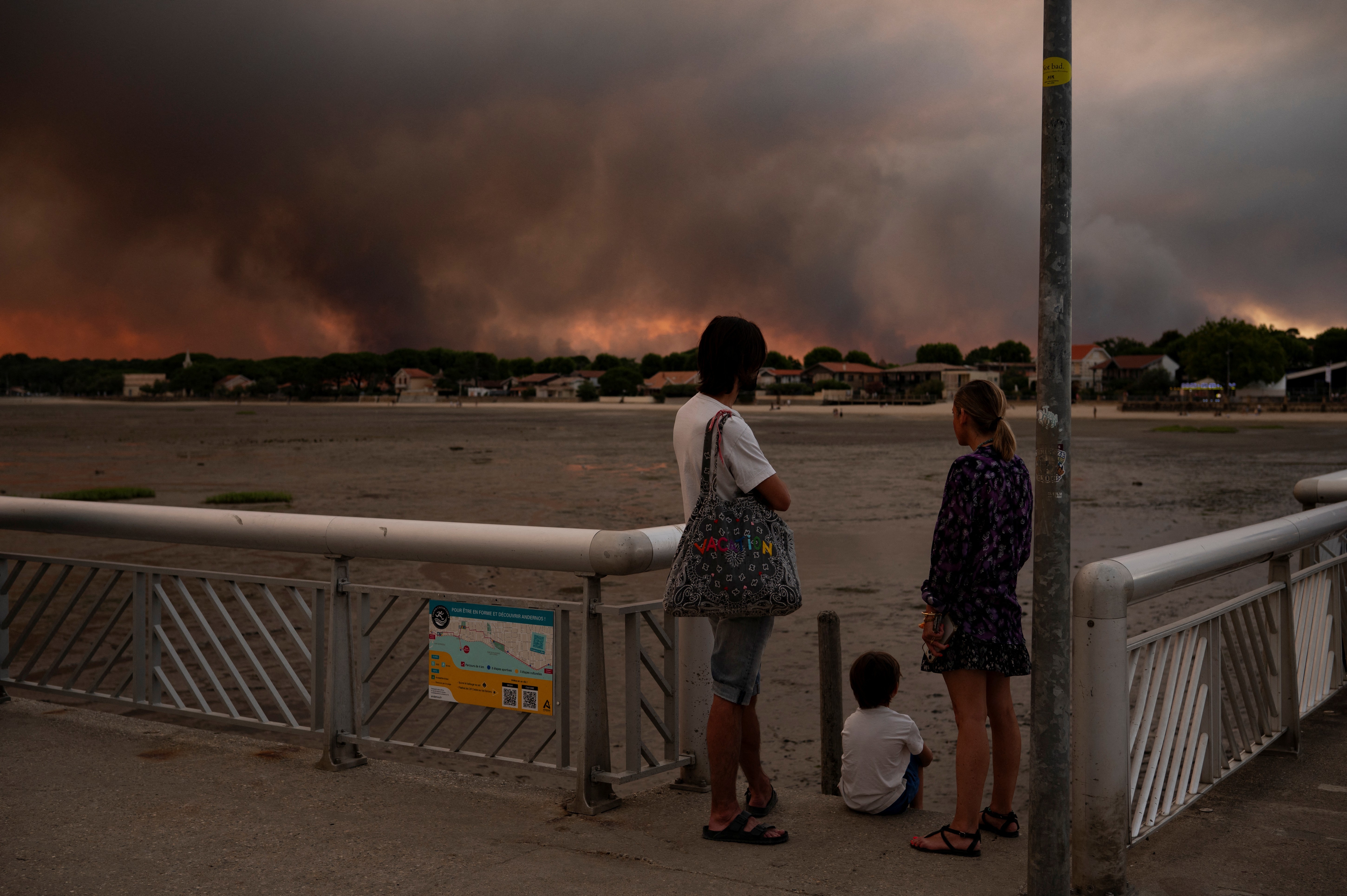 Uma família, de costas, observa uma densa fumaça escura no horizonte, com tons alaranjados do pôr do sol. Um adulto com camiseta branca e bolsa, uma mulher de vestido estampado e uma criança sentada no chão estão em um píer com corrimão metálico. Ao fundo, casas e vegetação sob o céu escurecido pela fumaça.