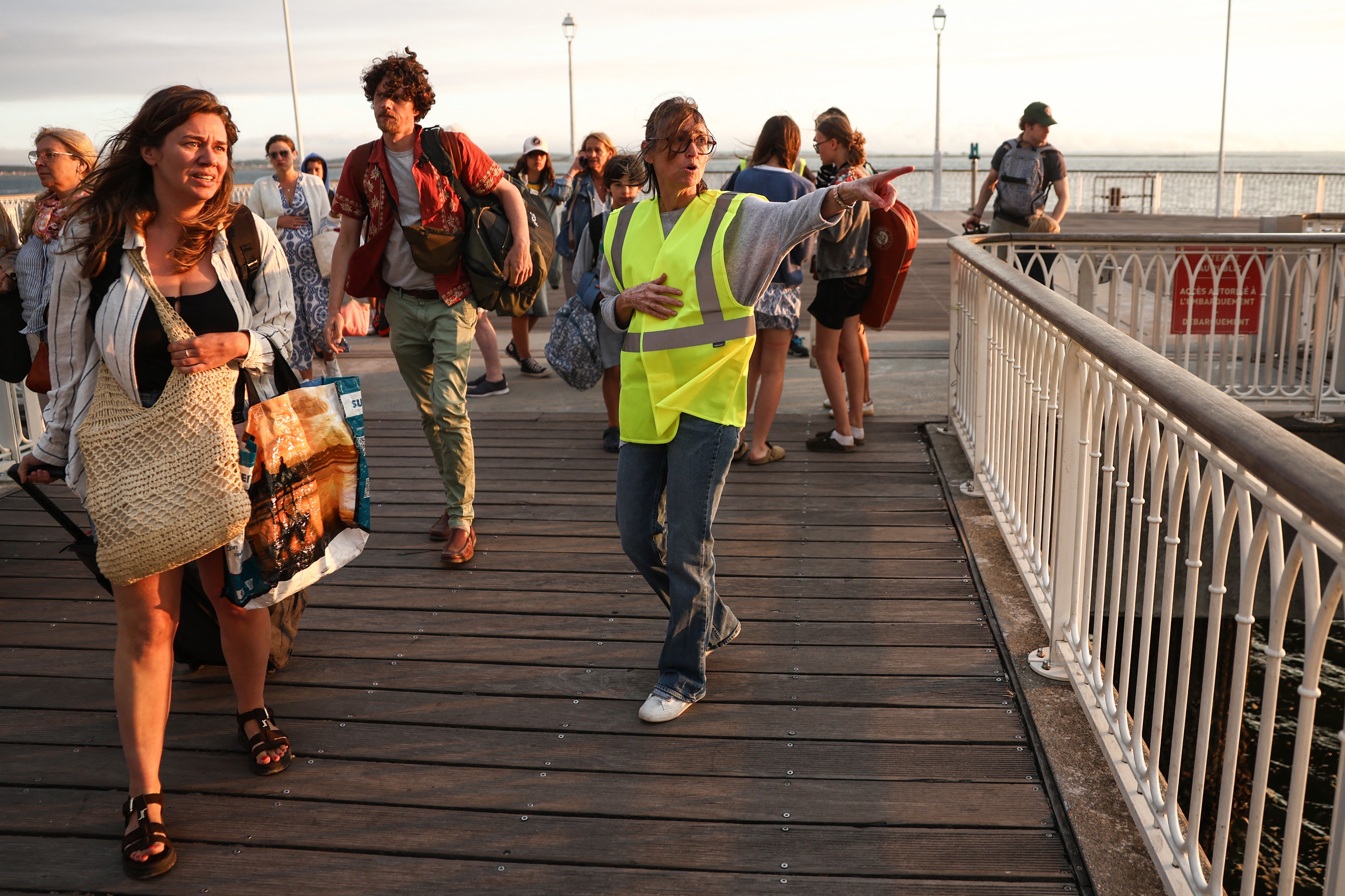 Pessoas caminhando em um píer de madeira ao pôr do sol. Uma mulher de colete amarelo fluorescente aponta para a direita, orientando um grupo de turistas. Outra mulher à esquerda carrega uma bolsa de palha e uma sacola grande, enquanto um homem com mochila caminha atrás dela. O corrimão branco do píer e o mar ao fundo completam a cena