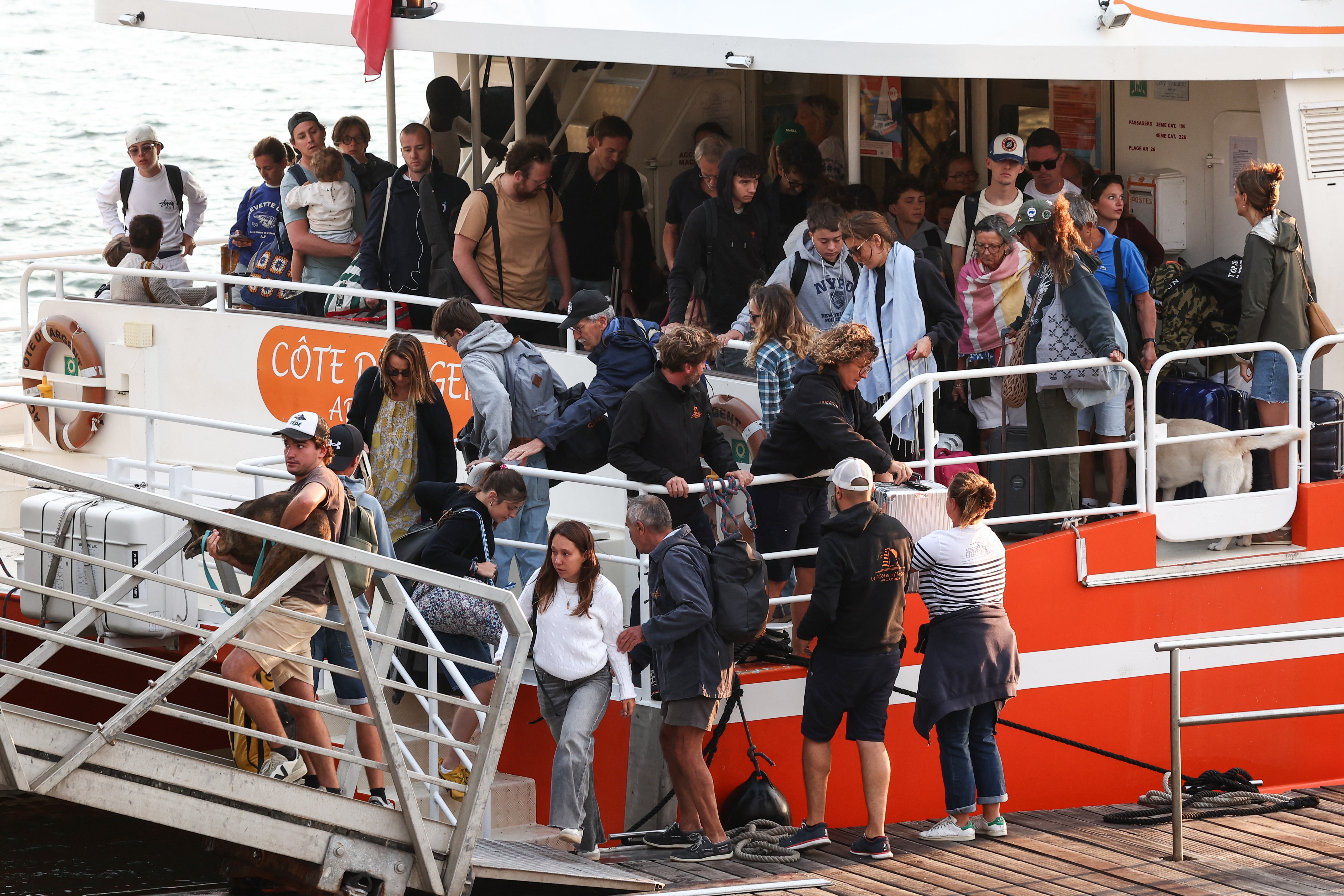 Pessoas de diversas idades desembarcam de um barco de passageiros branco e laranja, com a inscrição CÔTE DAZUR visível. Alguns carregam mochilas e malas, enquanto outros seguram crianças ou animais de estimação, como um cachorro labrador claro. A rampa de metal do barco conecta-se a um cais de madeira, e a cena sugere um dia movimentado de viagem ou passeio