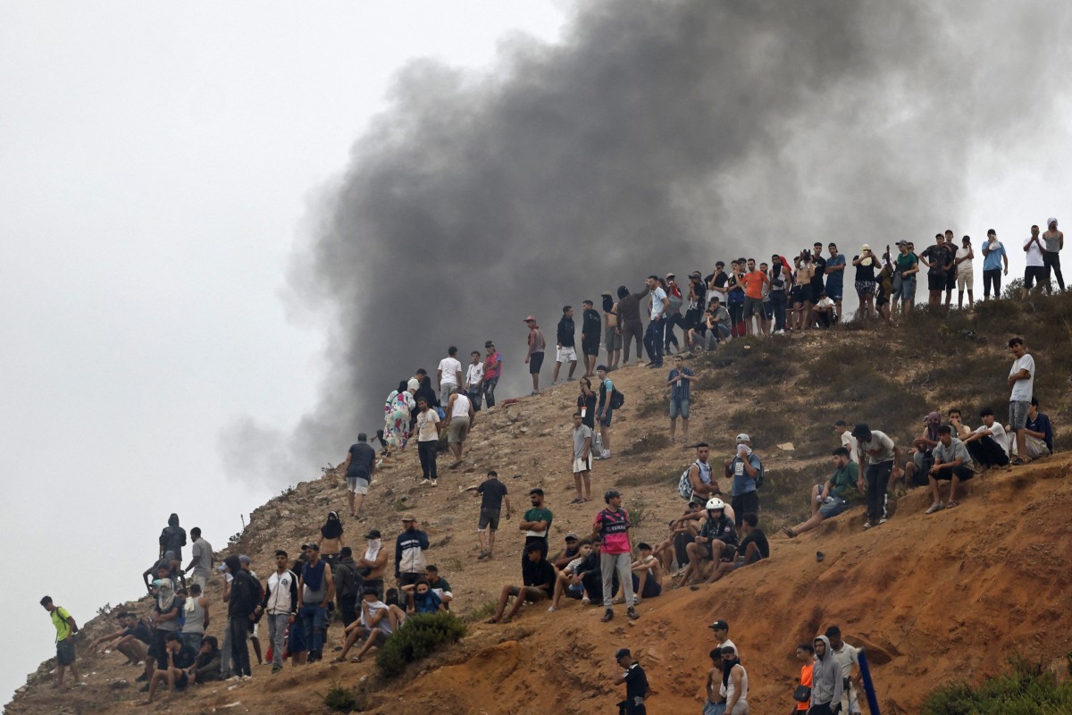 Multidão de pessoas, a maioria jovens, em uma encosta de terra marrom, observando uma densa fumaça escura que sobe no céu cinzento ao fundo