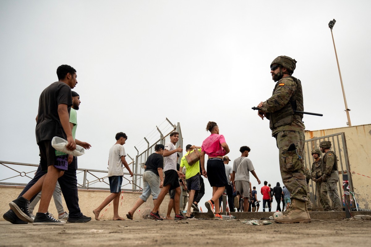 Grupo de jovens e homens caminhando em direção a uma cerca de arame farpado, enquanto um soldado com uniforme camuflado e óculos escuros os observa, segurando um bastão. Outros soldados estão ao fundo, perto de uma estrutura bege. O céu está nublado e claro.