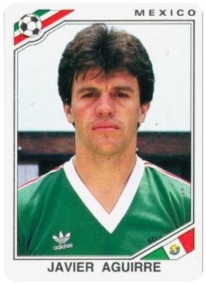 Cartão de figurinha do jogador de futebol Javier Aguirre, com cabelo castanho escuro e camisa verde com gola branca em V, olhando diretamente para frente. No canto superior esquerdo, uma bola de futebol e as cores da bandeira do México. No canto inferior direito, a bandeira do México. No topo, a palavra MEXICO e na parte inferior JAVIER AGUIRRE